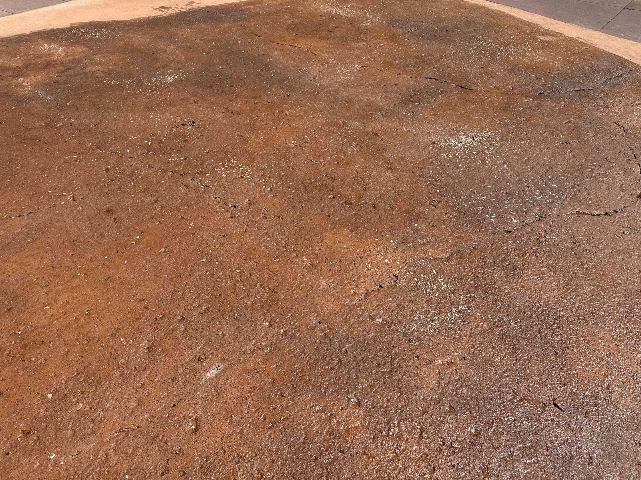 A flat, reddish-brown concrete surface with visible cracks and light speckles under bright sunlight.