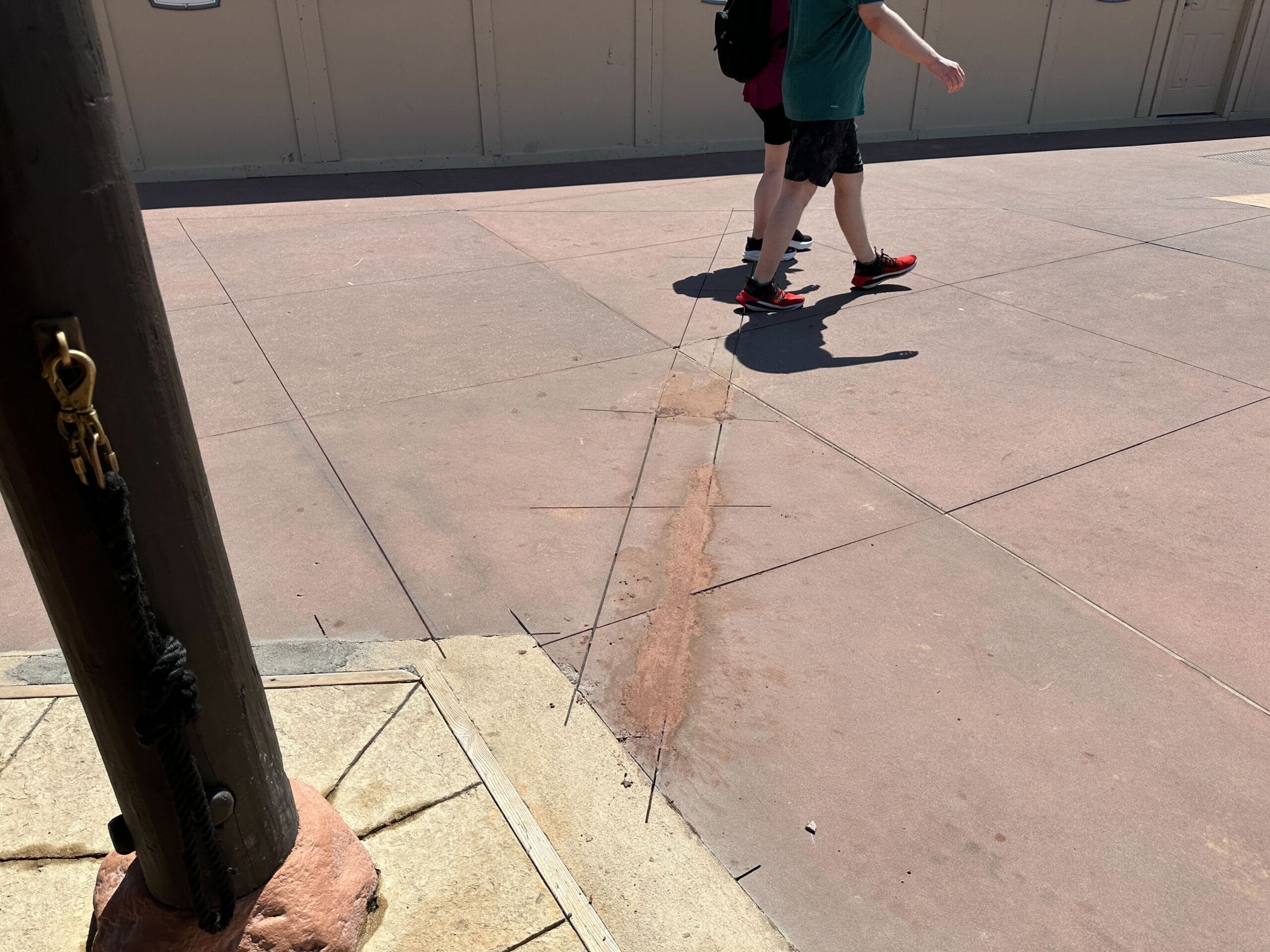 A person walks on a sunlit sidewalk with a long, rust-colored stain running across the pavement.