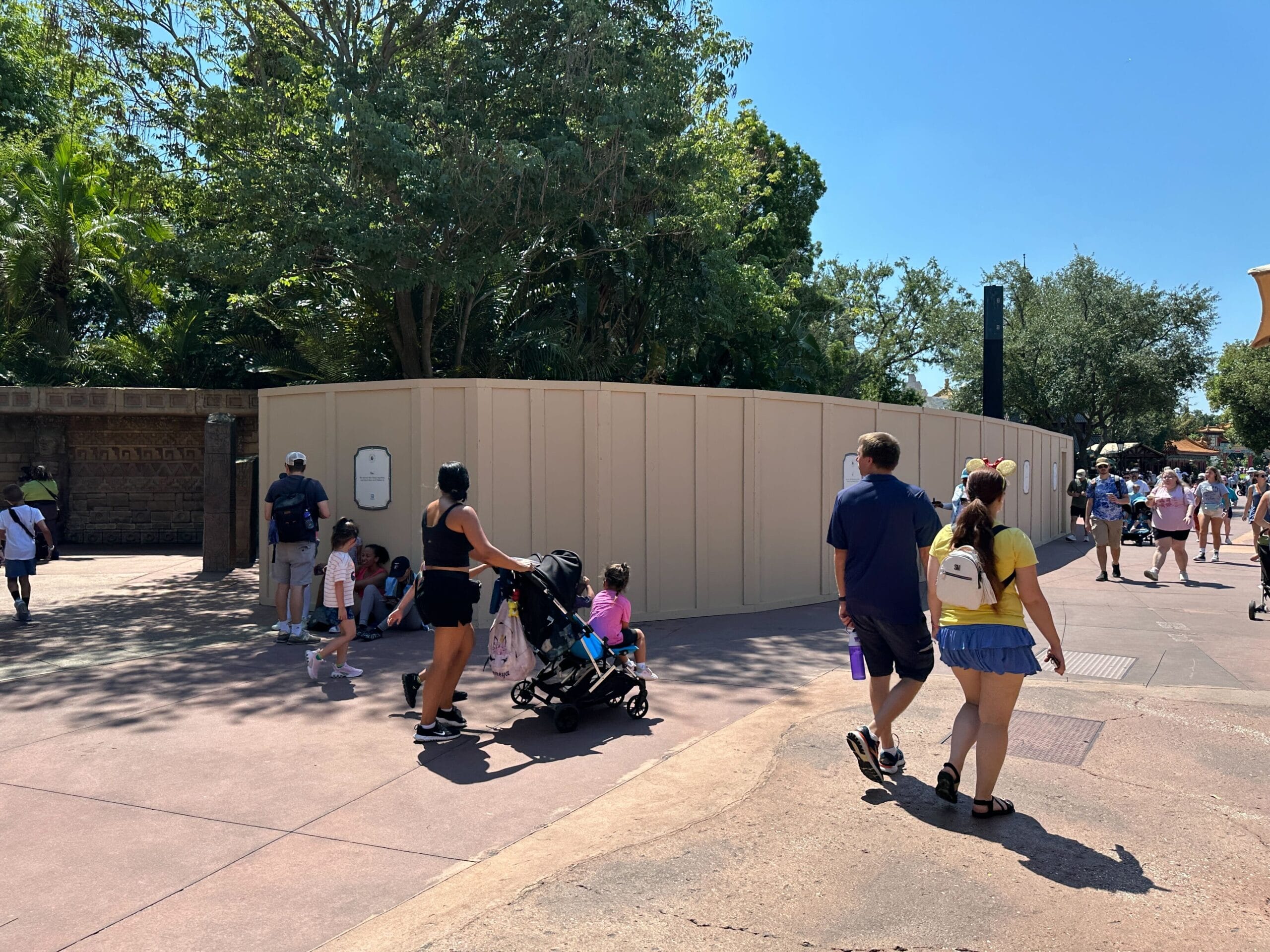 People walk past a beige construction wall at a theme park on a sunny day, with some sitting by the wall.
