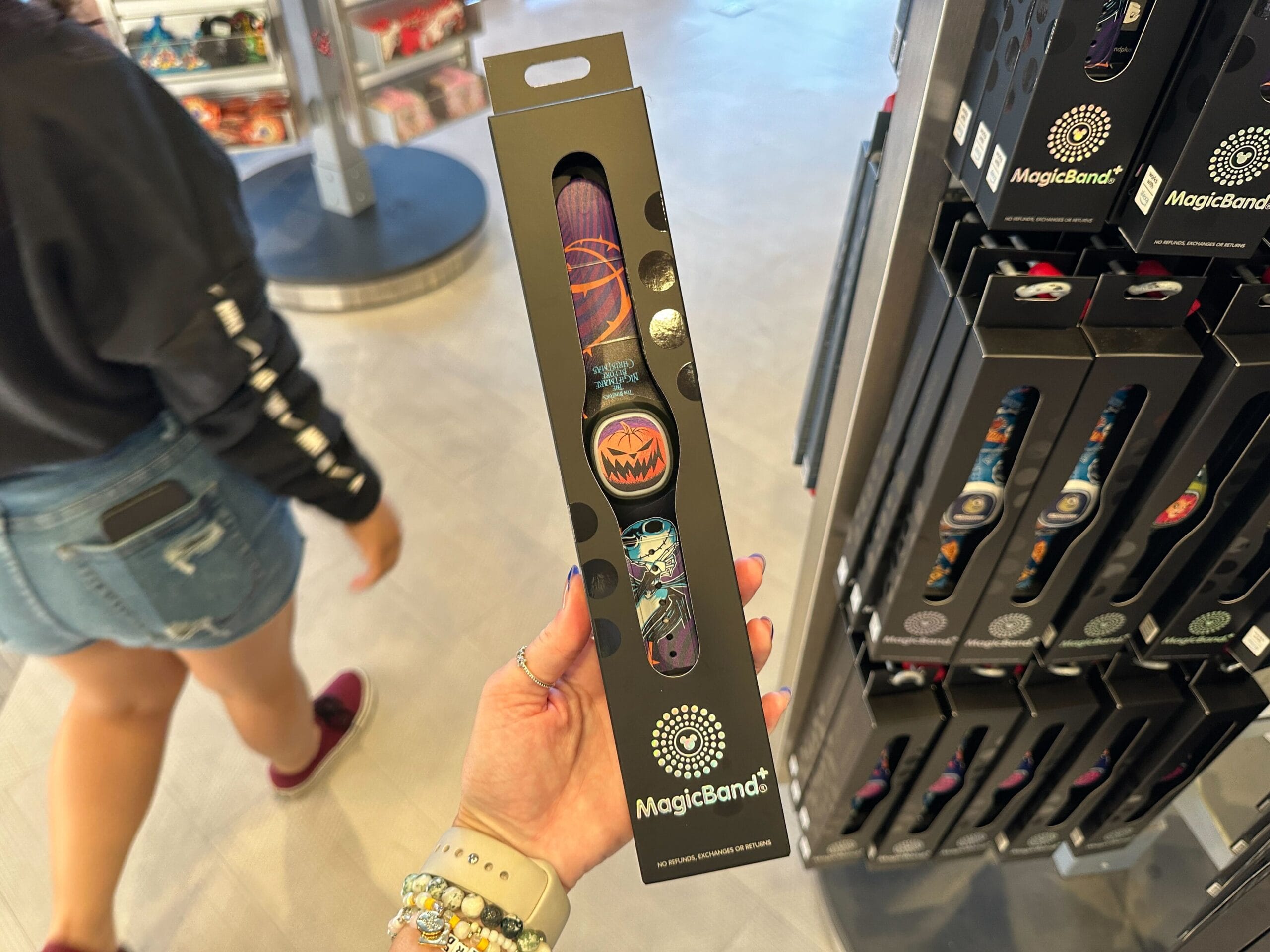 Person holds up a boxed MagicBand+ with Nightmare Before Christmas designs in an EPCOT store; other bands visible.