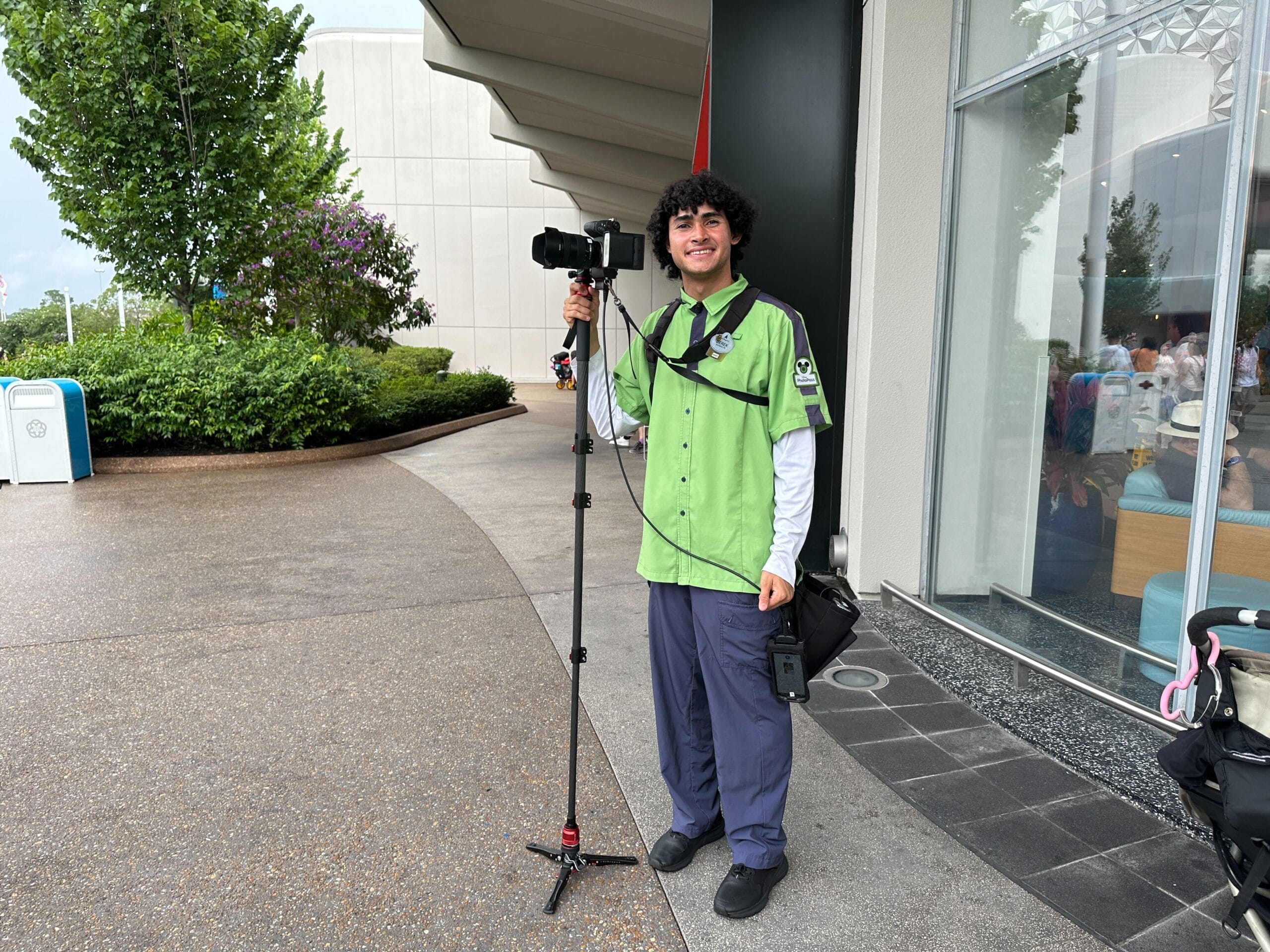 A smiling man in a green shirt stands outside holding a camera on a tripod near a modern building.