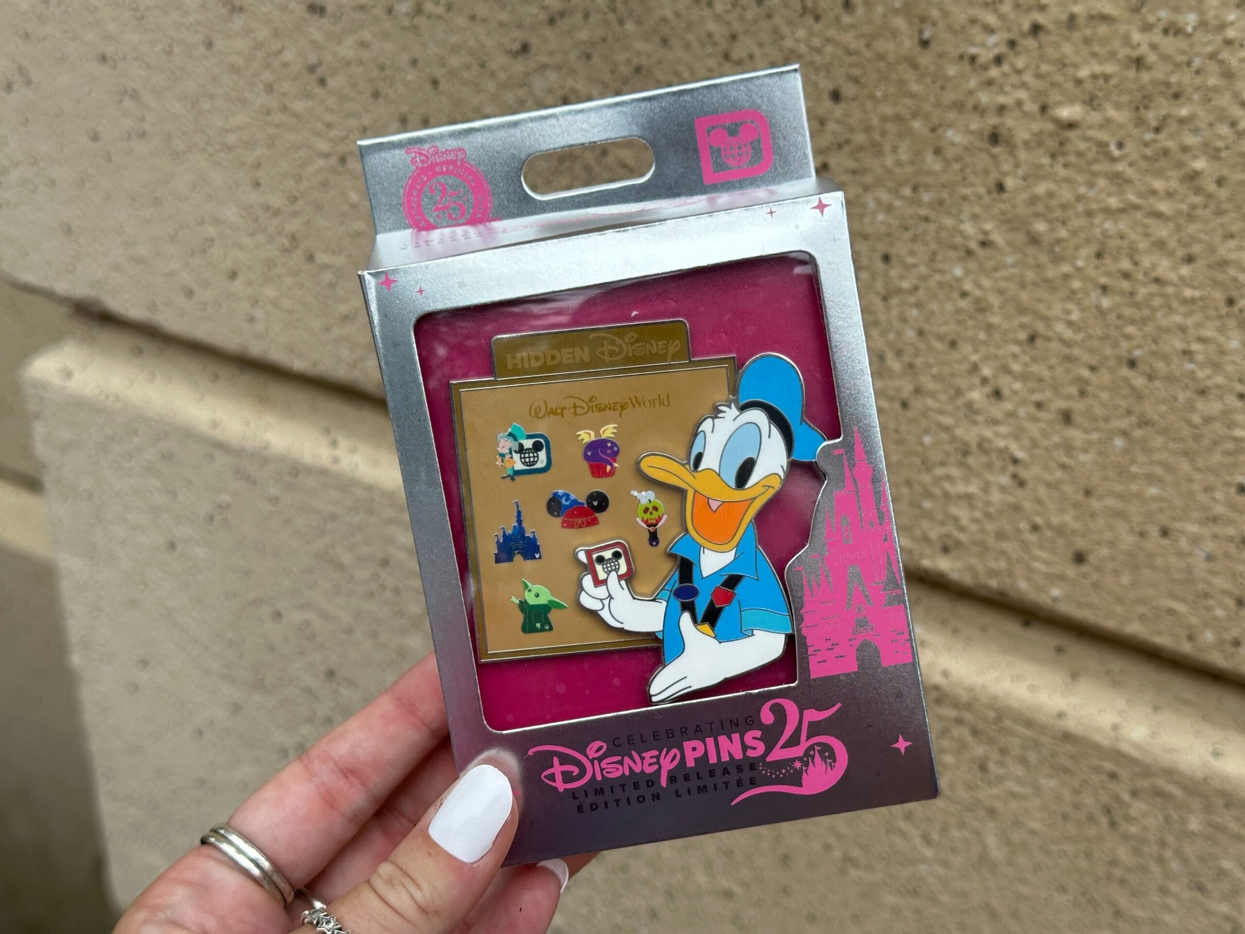 Hand holding a Disney Pins box featuring Donald Duck and colorful Disney park pins on a pink and silver background.