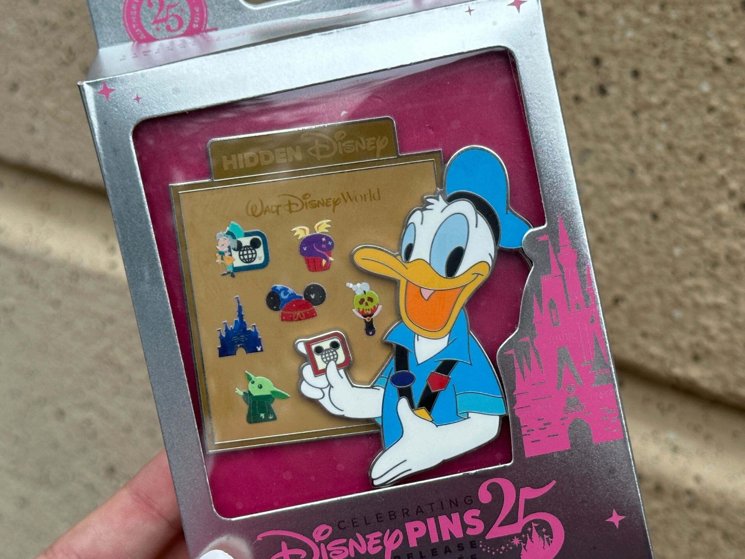 A hand holds a Disney pin set featuring Donald Duck and various icons in a 25th anniversary package.