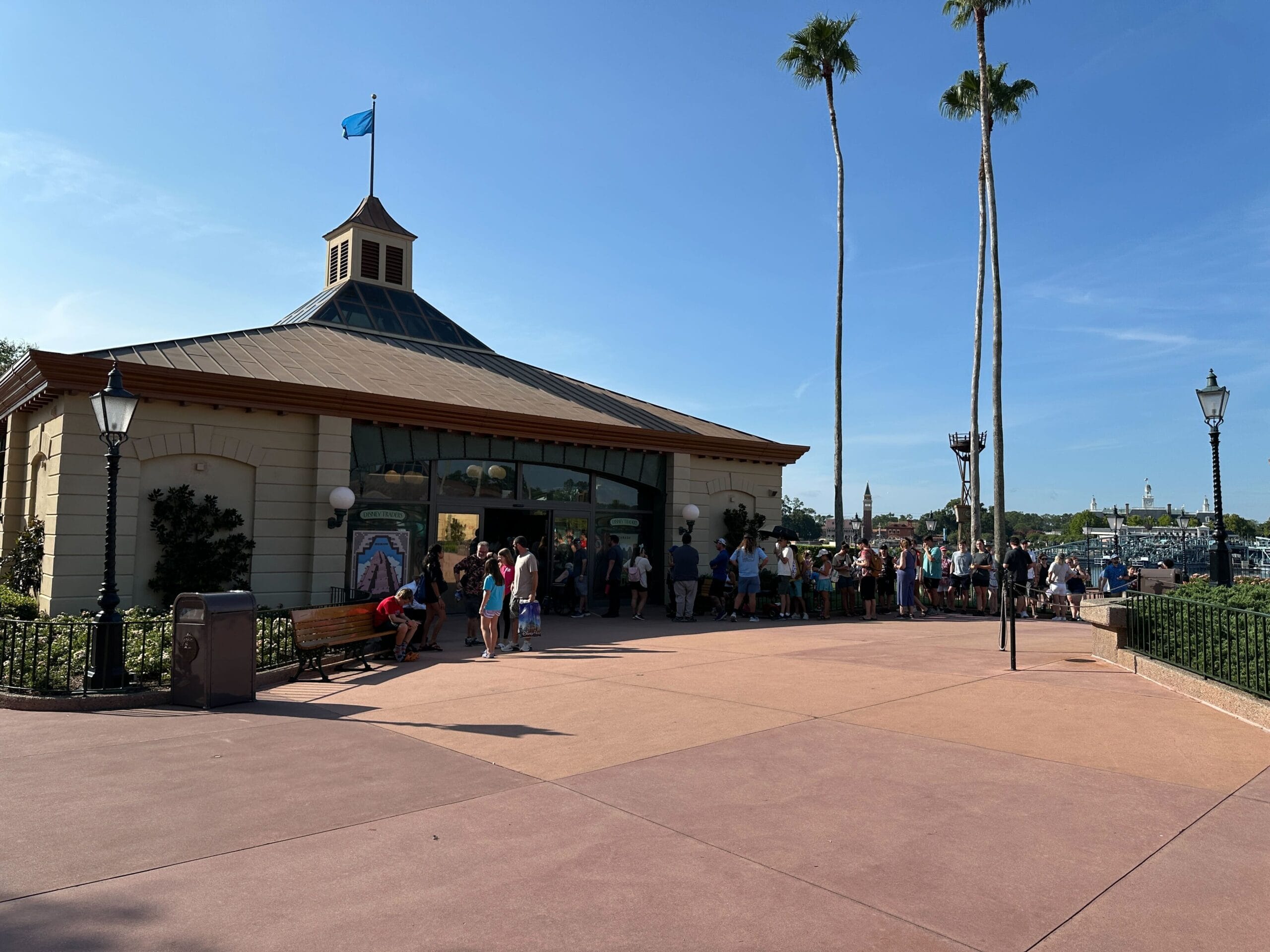 A group of people waits in line outside a beige building with a blue flag on a clear, sunny day.