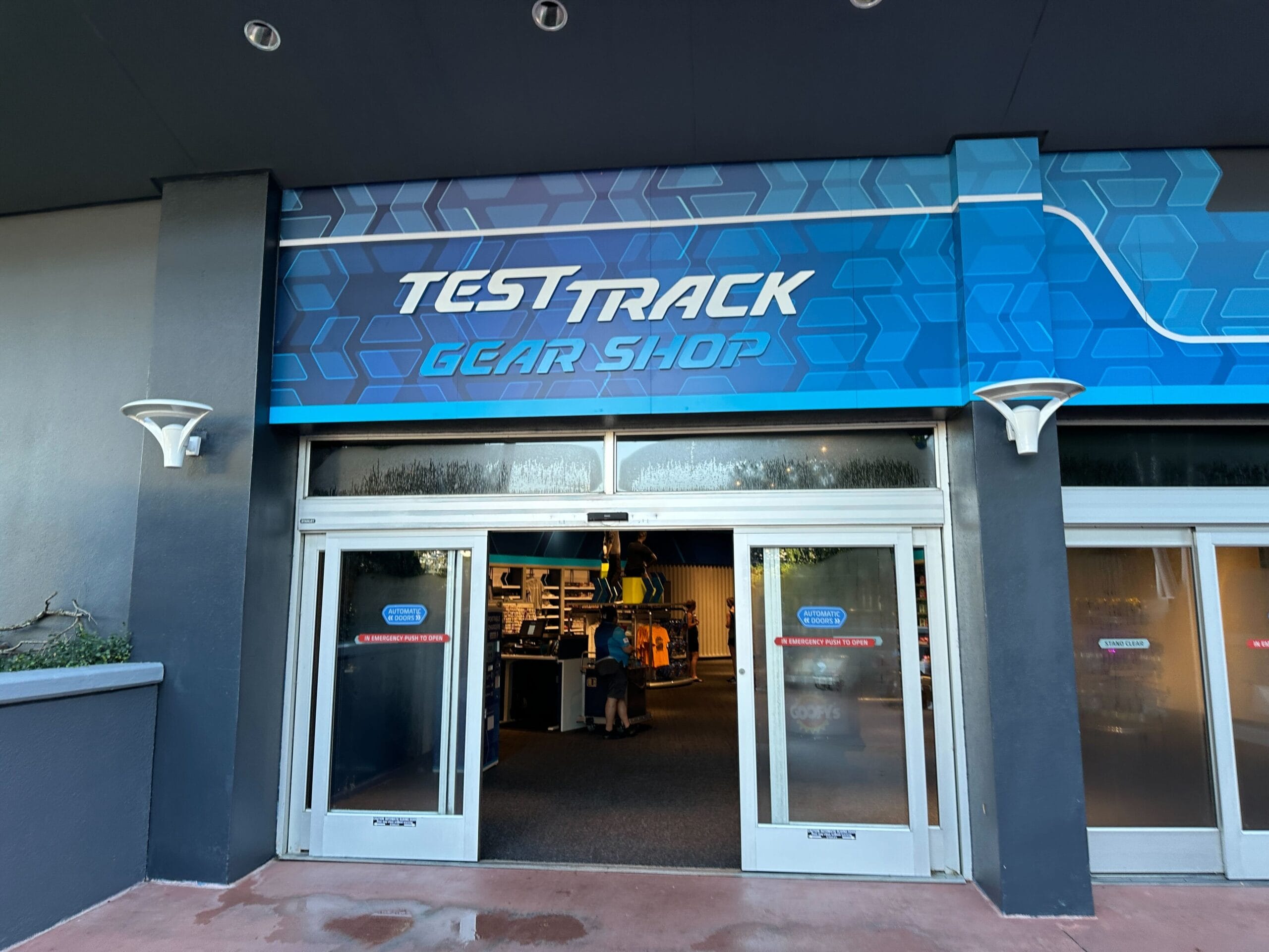 Entrance to the EPCOT Test Track Gear Shop with automatic glass doors and a blue geometric sign above.