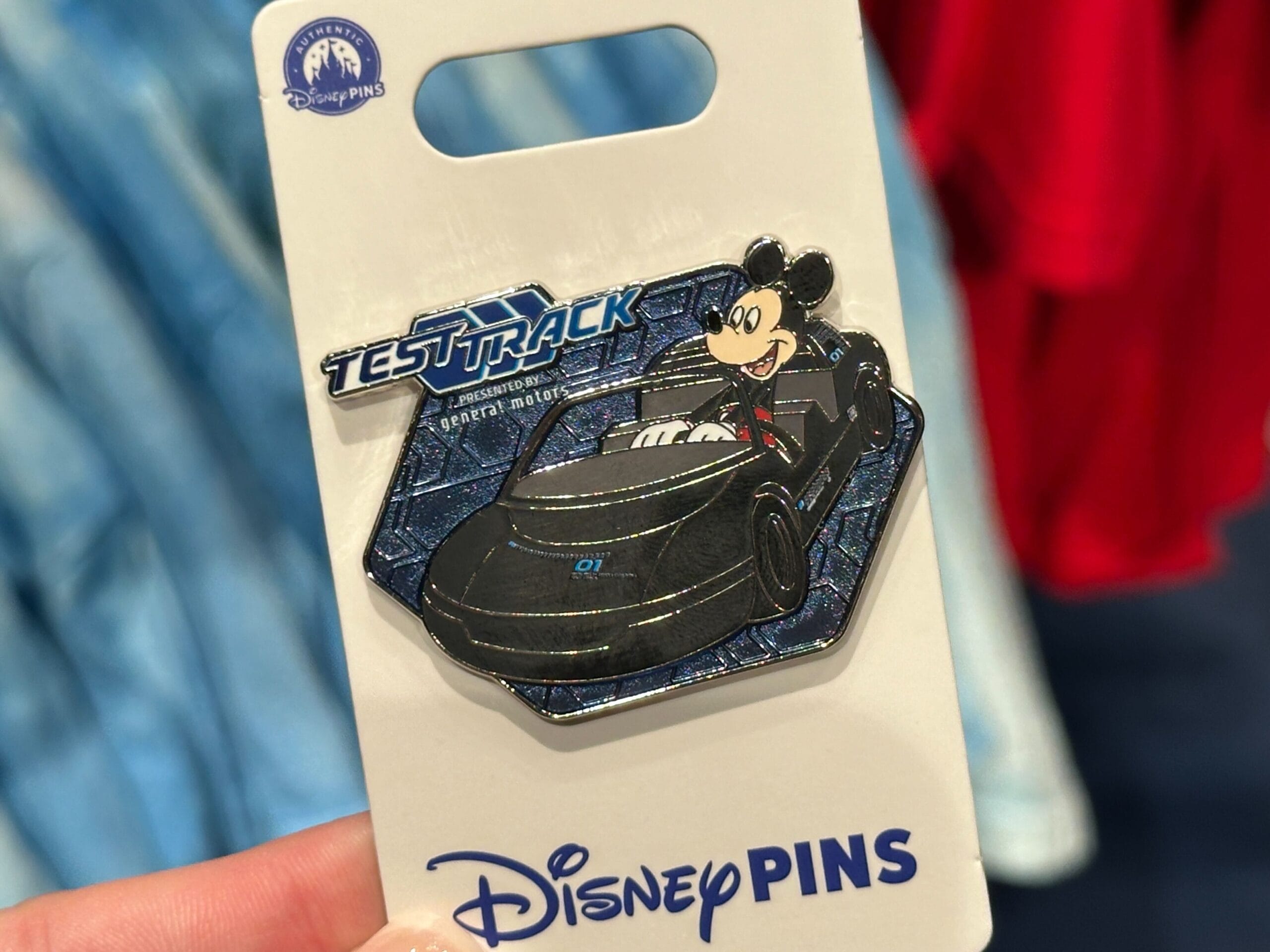 A hand holds a Disney Test Track pin from EPCOT, featuring Mickey Mouse driving a futuristic black car on a display card.
