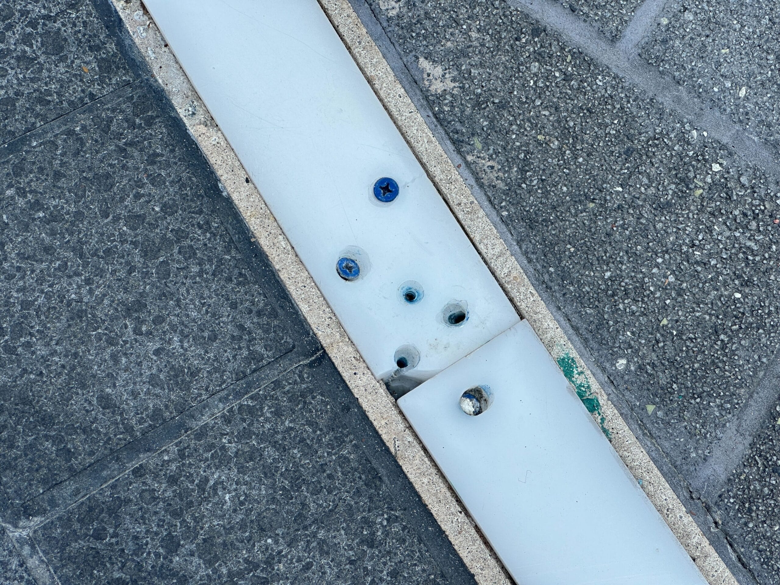 Close-up of a sidewalk with a white panel, blue screws, and empty holes along the edge of textured gray tiles.