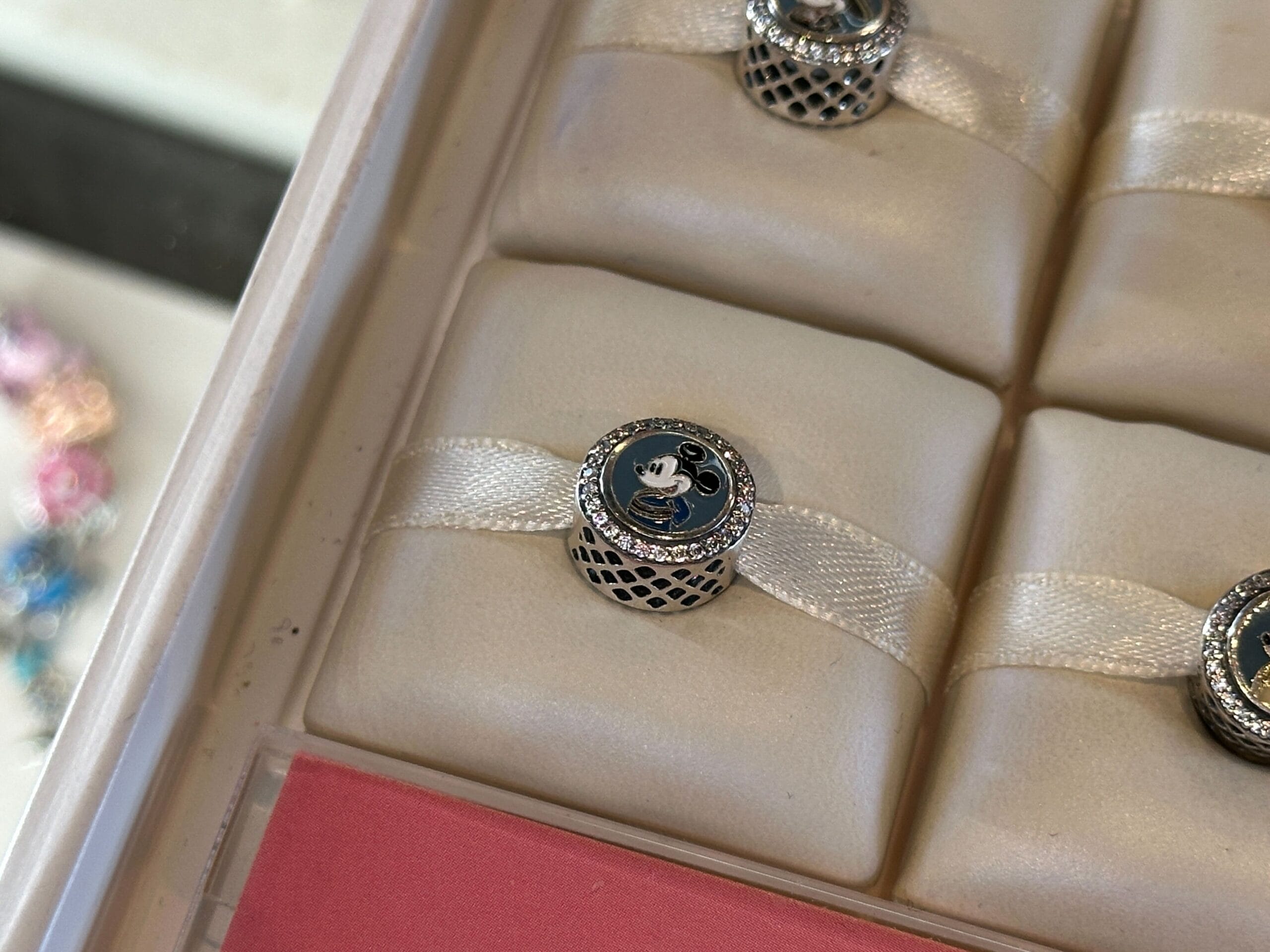 A round charm featuring a cartoon mouse face, displayed on a white ribbon in a jewelry case.