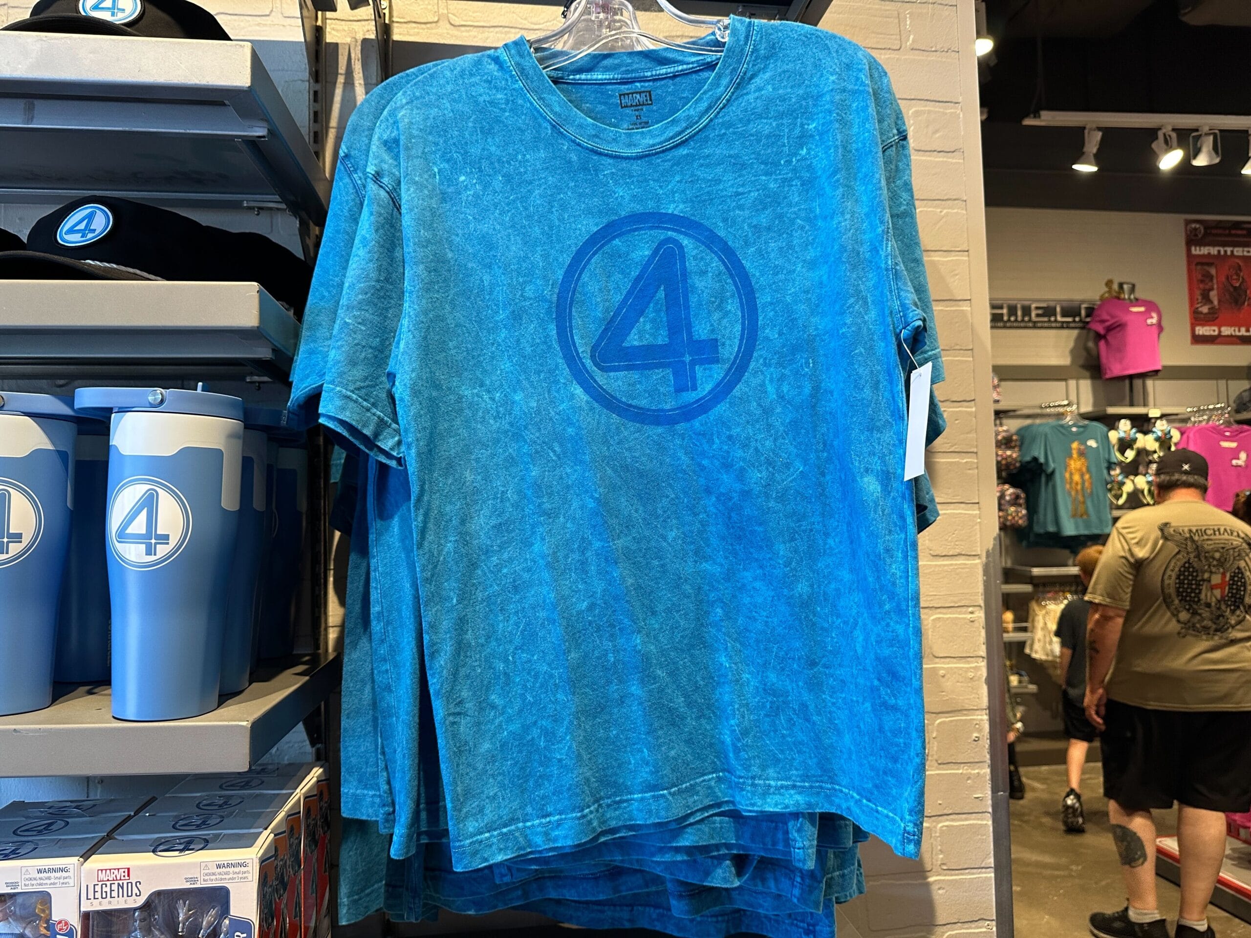 A blue Fantastic Four apparel T-shirt with the number 4 logo hangs on display in a comic store.