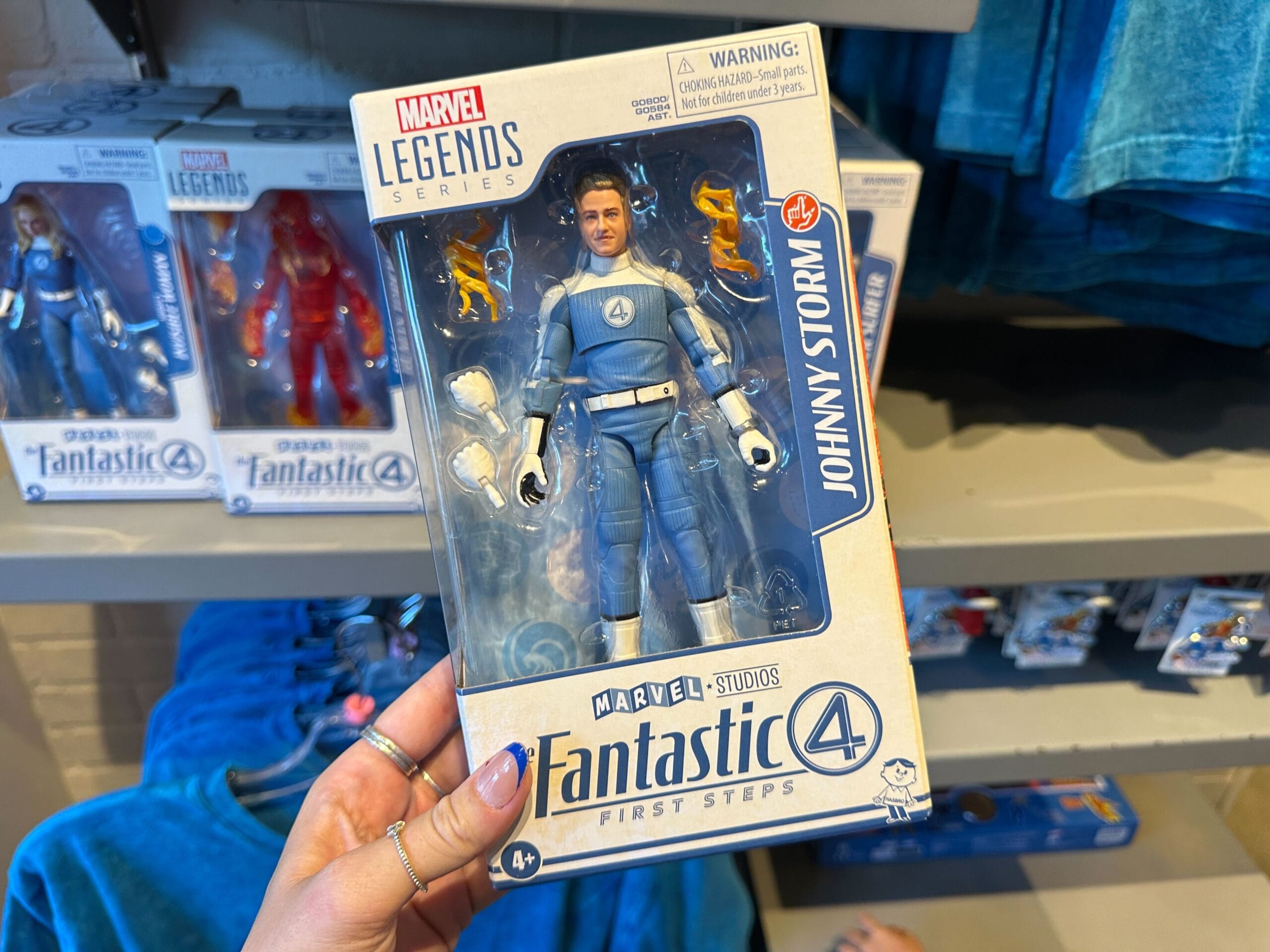 A hand holds a Marvel Legends Johnny Storm figure in blue, among Marvel merchandise at a store display.