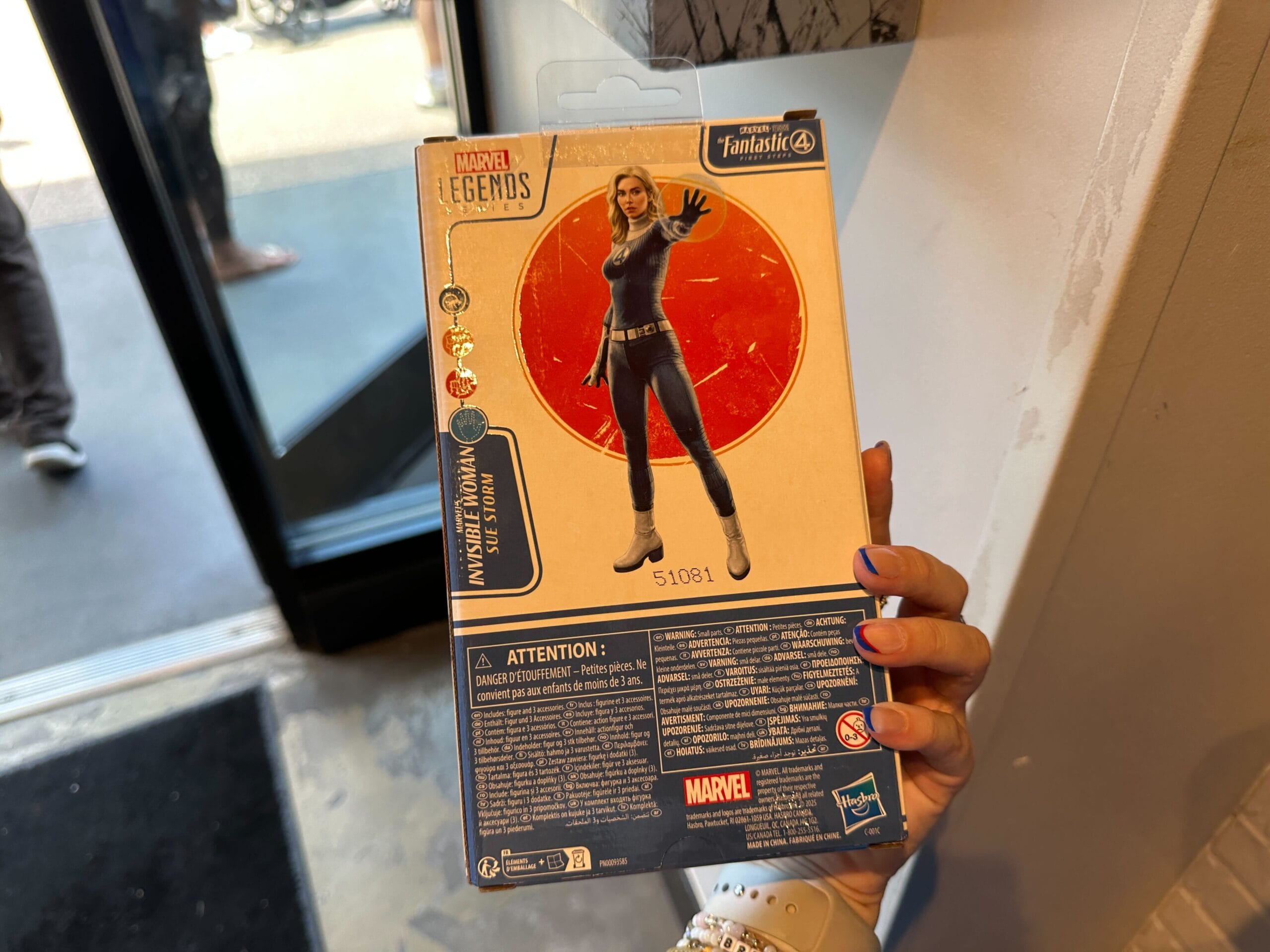 A hand holds Marvel merchandise—an Invisible Woman figure box—in a store near an open door.