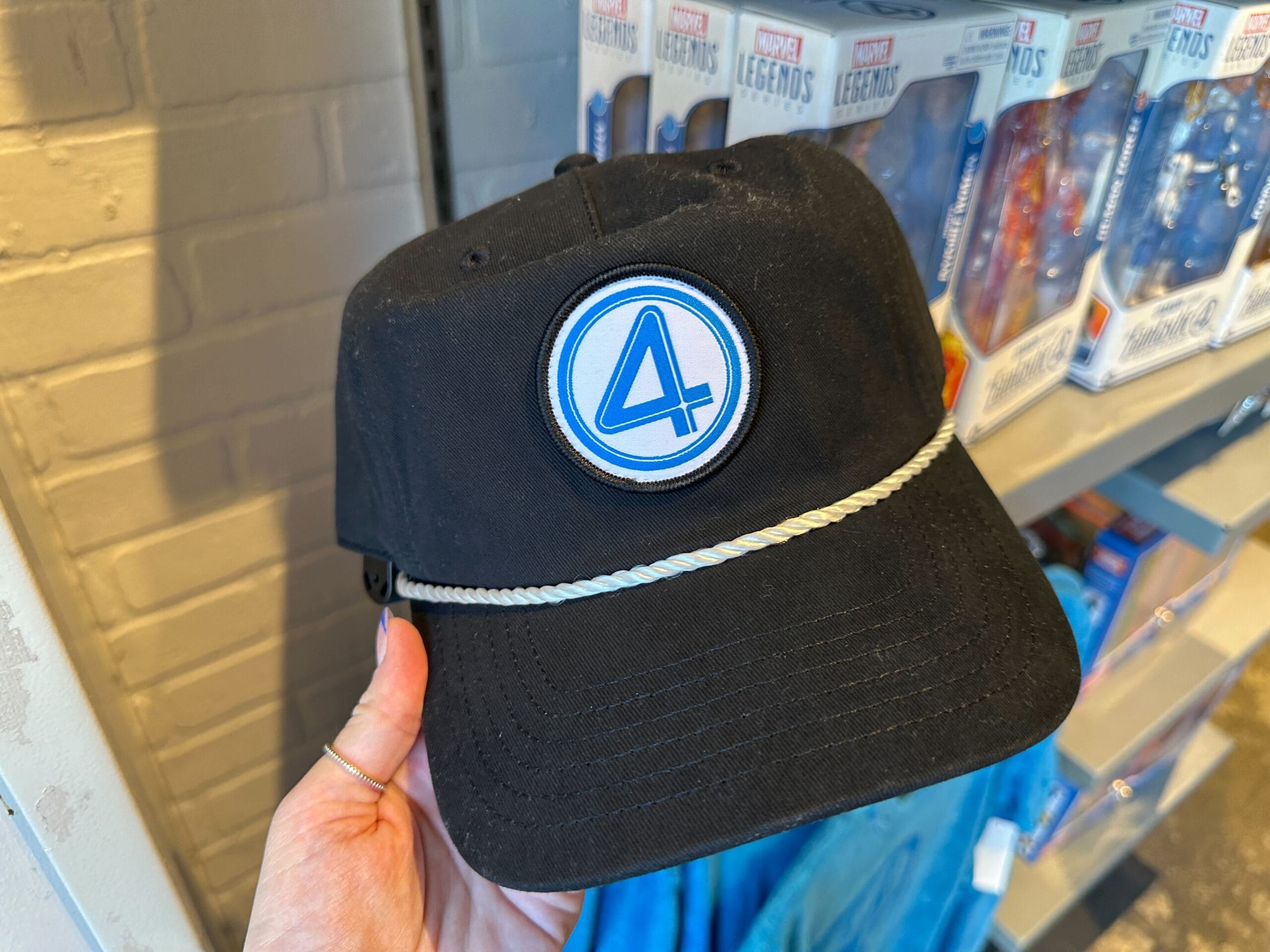 A hand holds Fantastic Four apparel—a black cap with a blue "4" logo, displayed near Marvel Legends toys in-store.