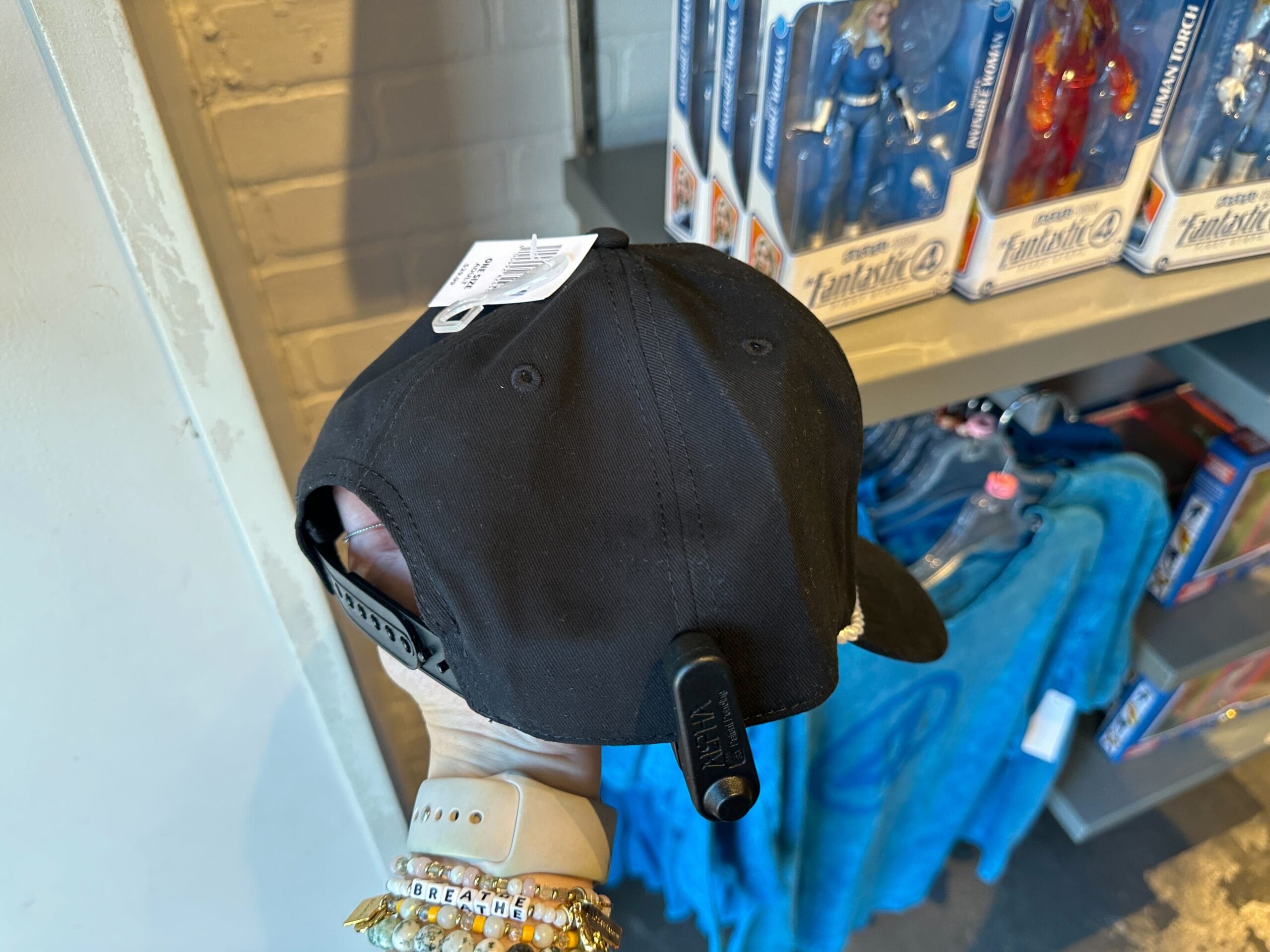 A hand holds a black cap with a security tag near Marvel Merchandise and Fantastic Four Apparel in the store.