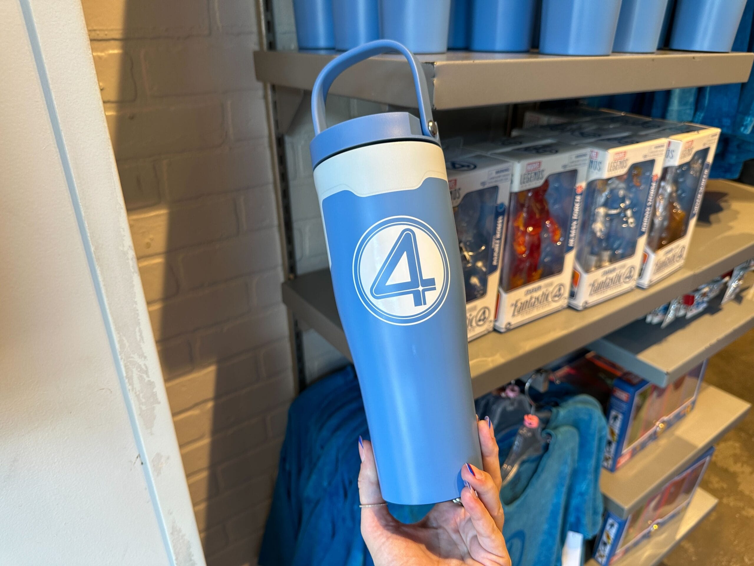 A hand holds a blue water bottle with a "4" logo in a store, surrounded by Fantastic Four apparel on the shelves behind.