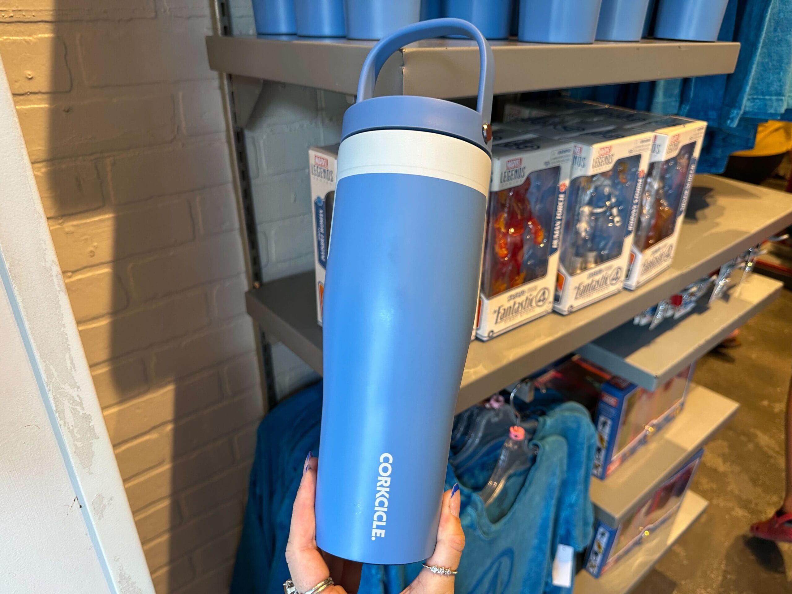A person holds a light blue Corkcicle water bottle in a store with Marvel merchandise on shelves in the background.
