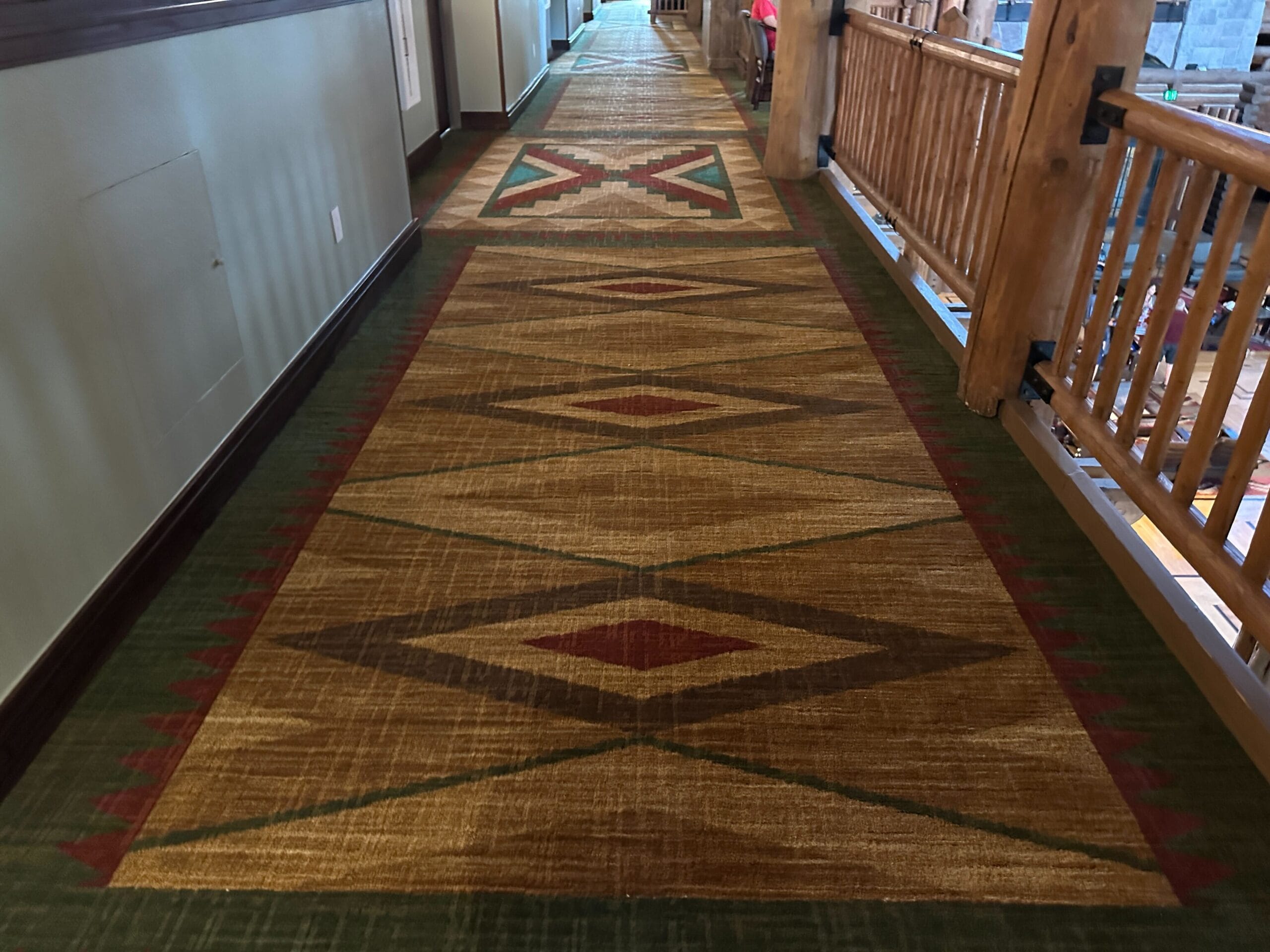 Hallway with a patterned carpet featuring diamond shapes in brown, red, and green, next to a wooden railing.