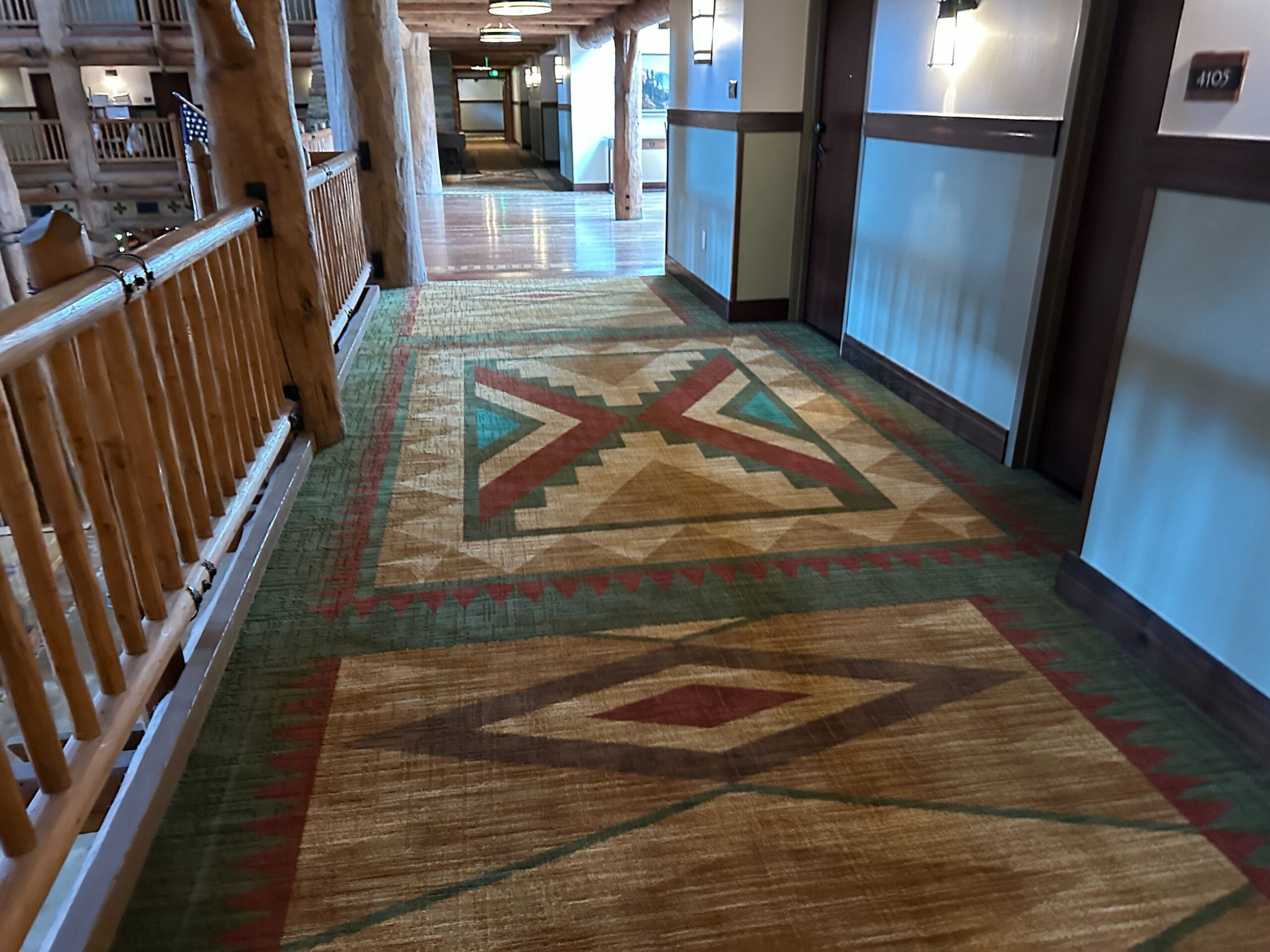 Hallway with patterned carpet, wooden railings, and doors to guest rooms in a rustic lodge-style building.