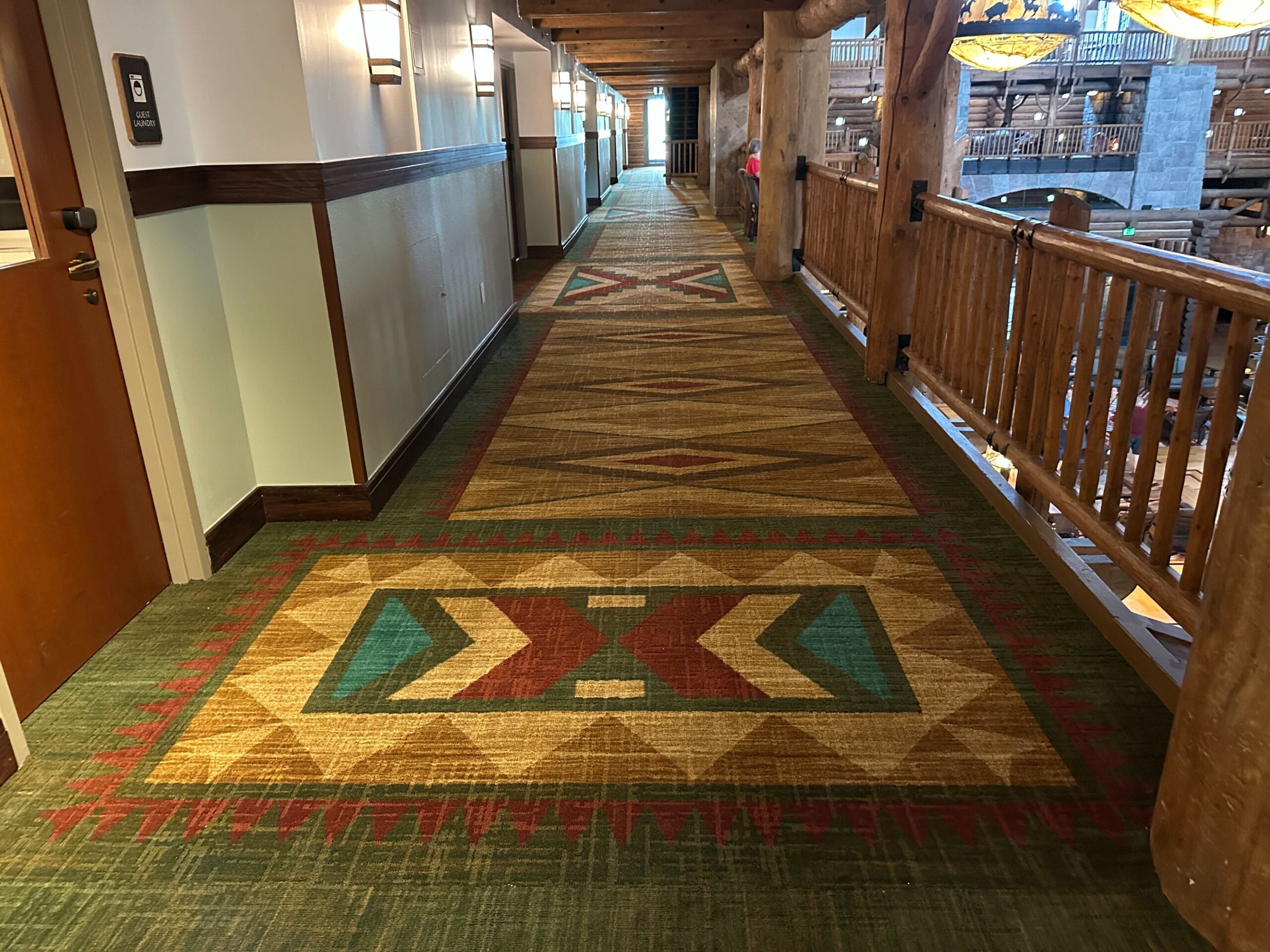 Hotel hallway with patterned carpet, wooden railings, and wall lamps, overlooking a lobby area on the right.