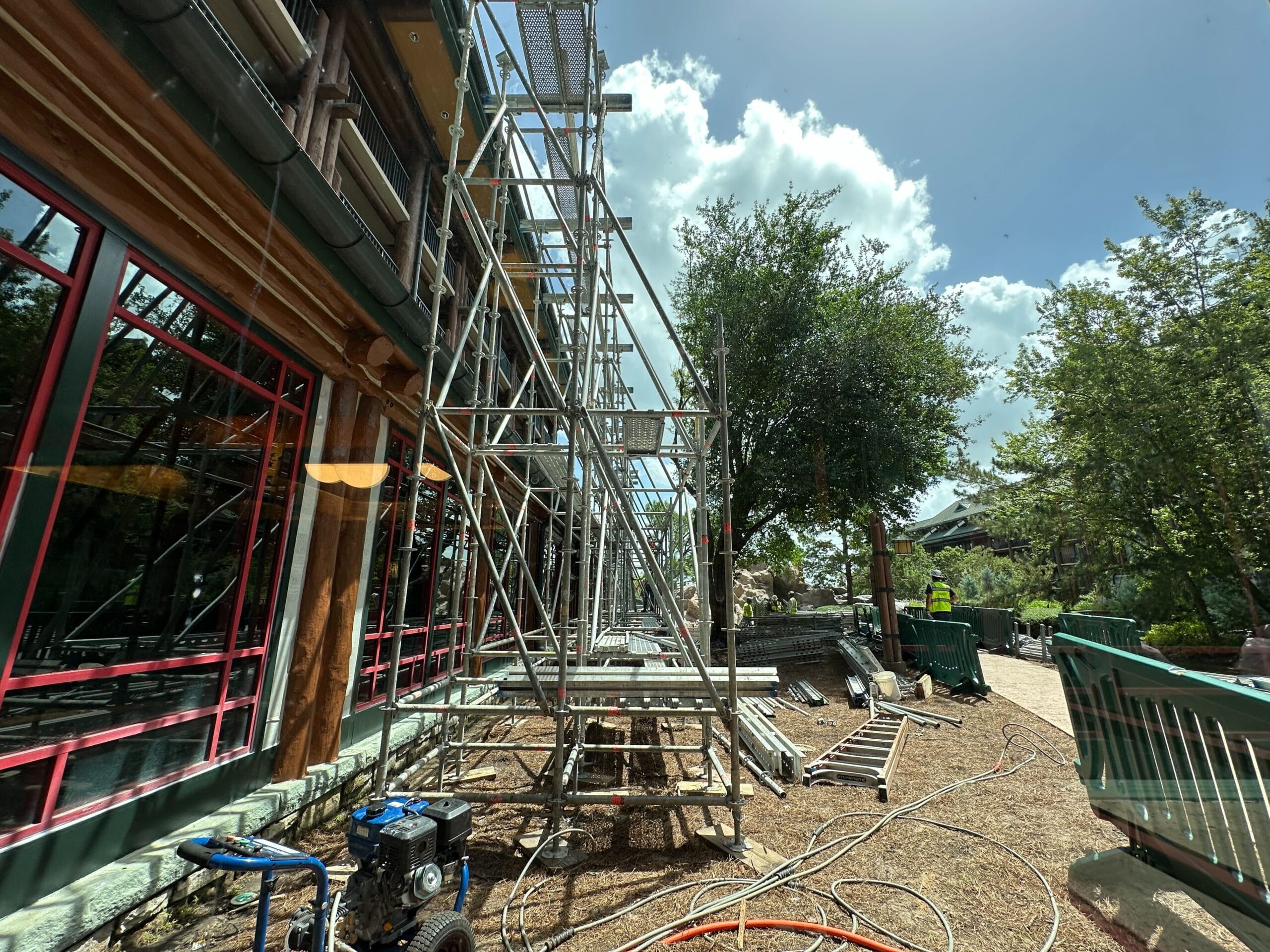 Scaffolding set up next to a building under a sunny sky, with trees and construction equipment nearby.