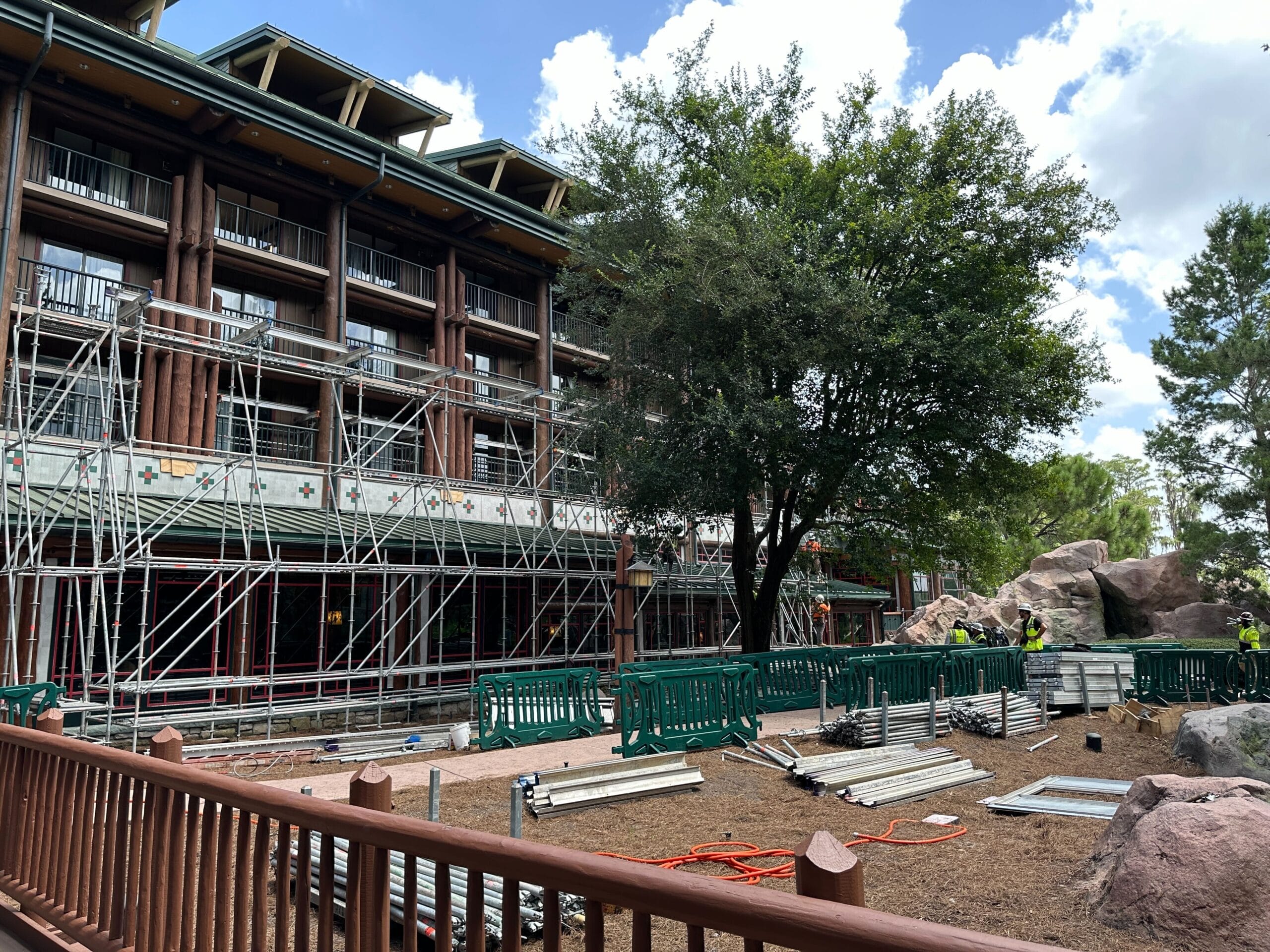 A building under construction with scaffolding, fencing, materials, and workers near trees on a partly cloudy day.