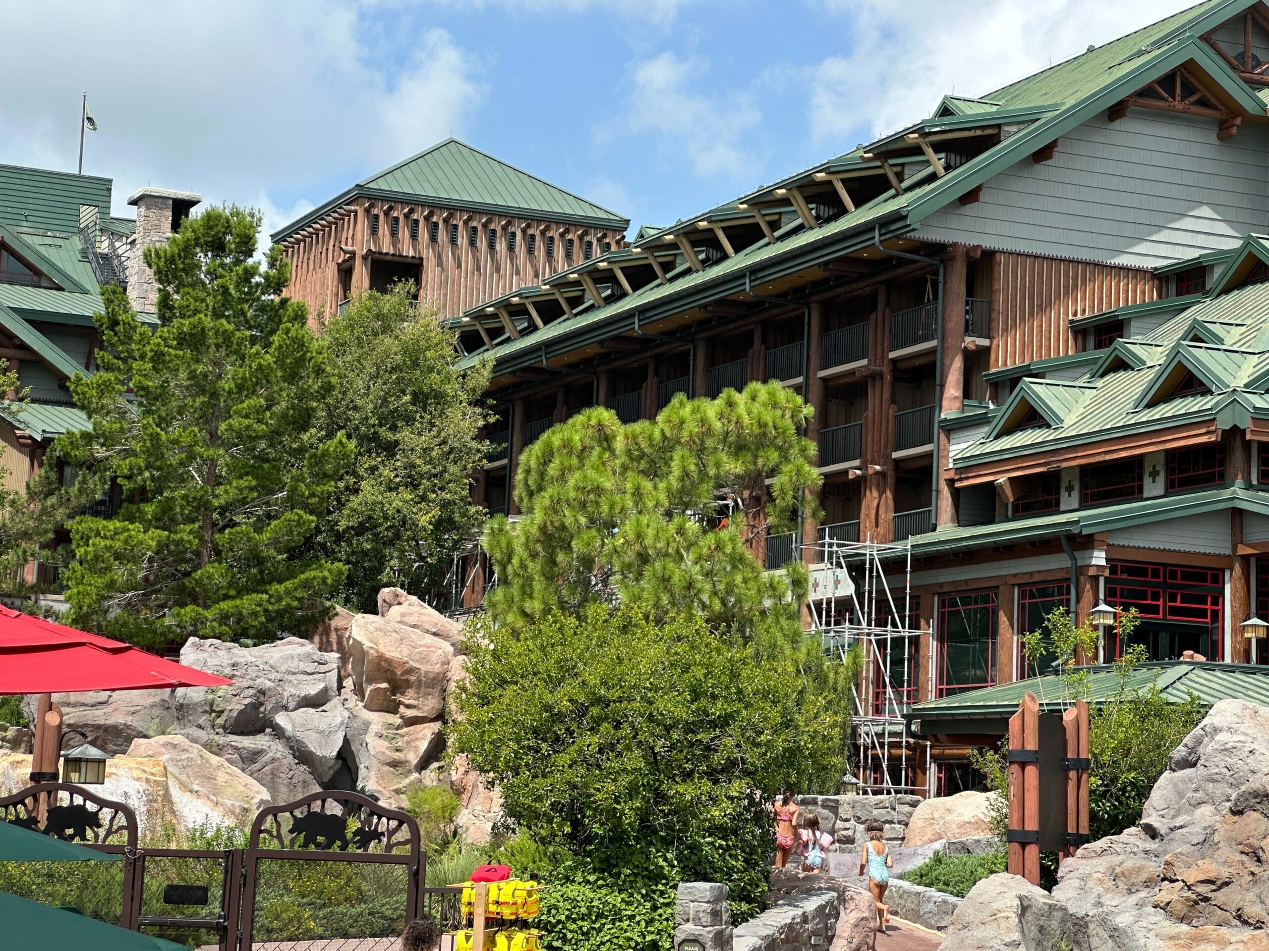 A rustic lodge with green roofs and scaffolding, surrounded by trees, rocks, and people walking nearby under a blue sky.
