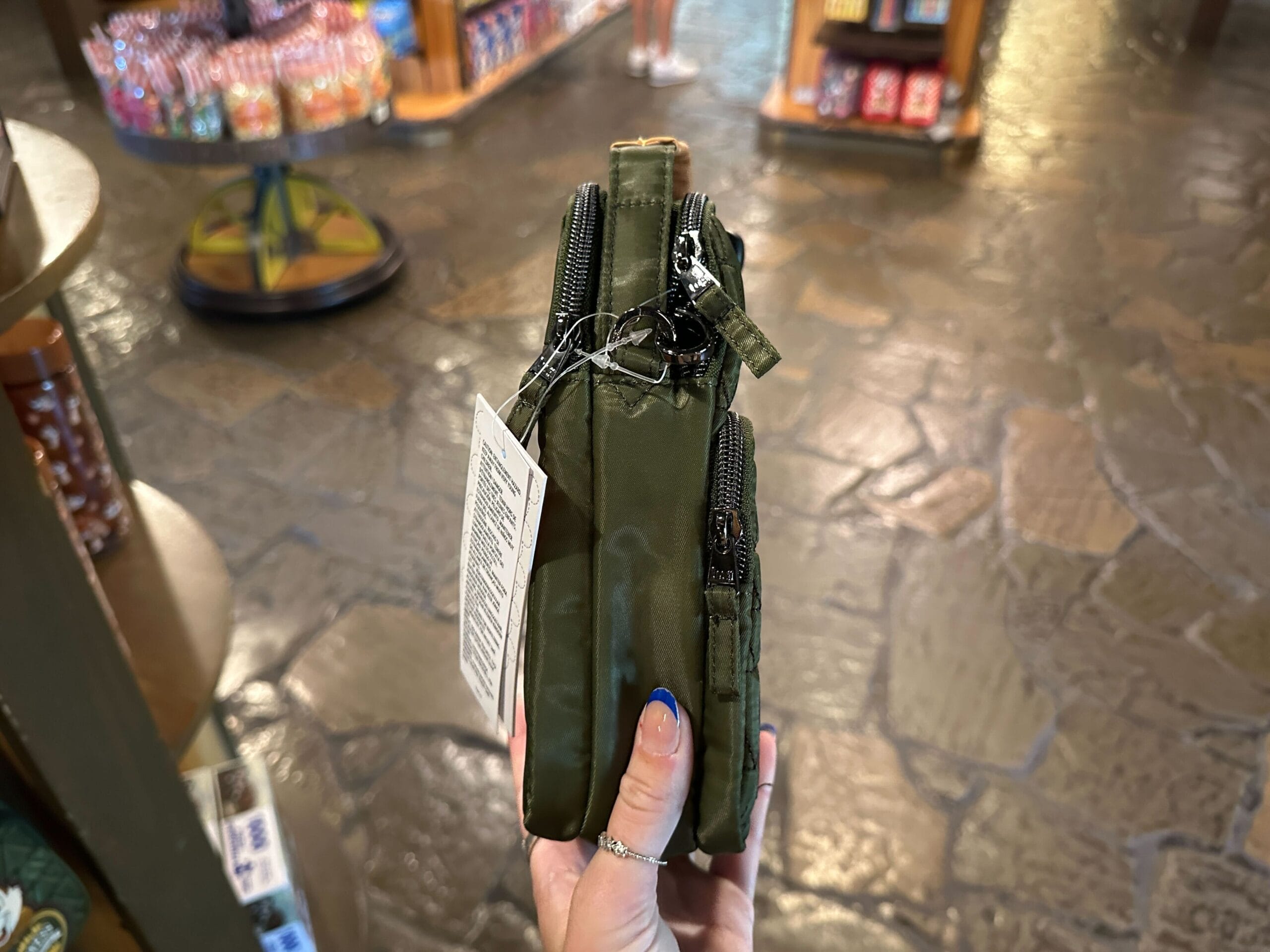 A hand holds a small green zippered pouch with tags in a store with stone flooring and shelves in the background.