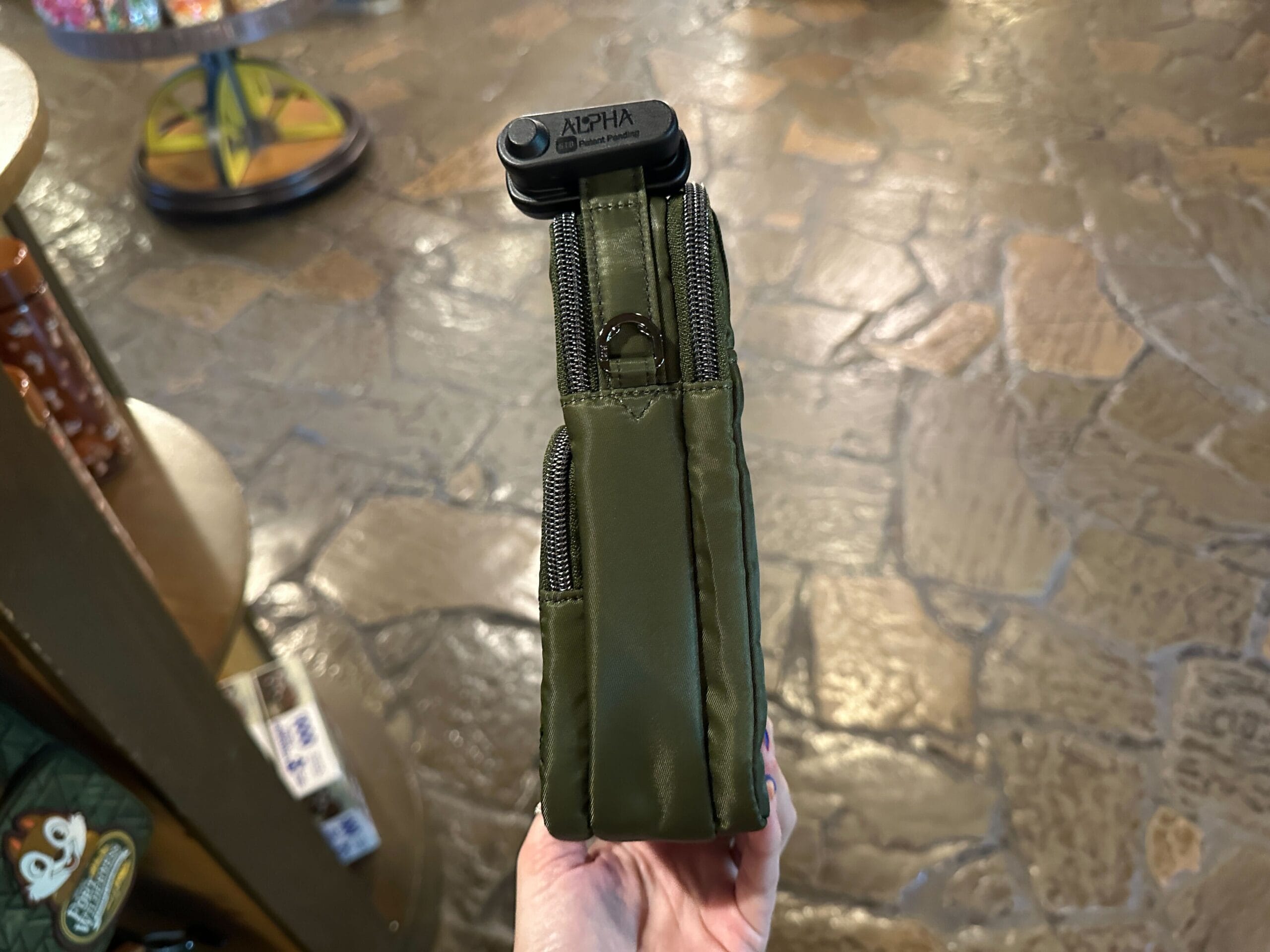 A hand holds an olive green zippered pouch upright in a store with stone-patterned flooring.