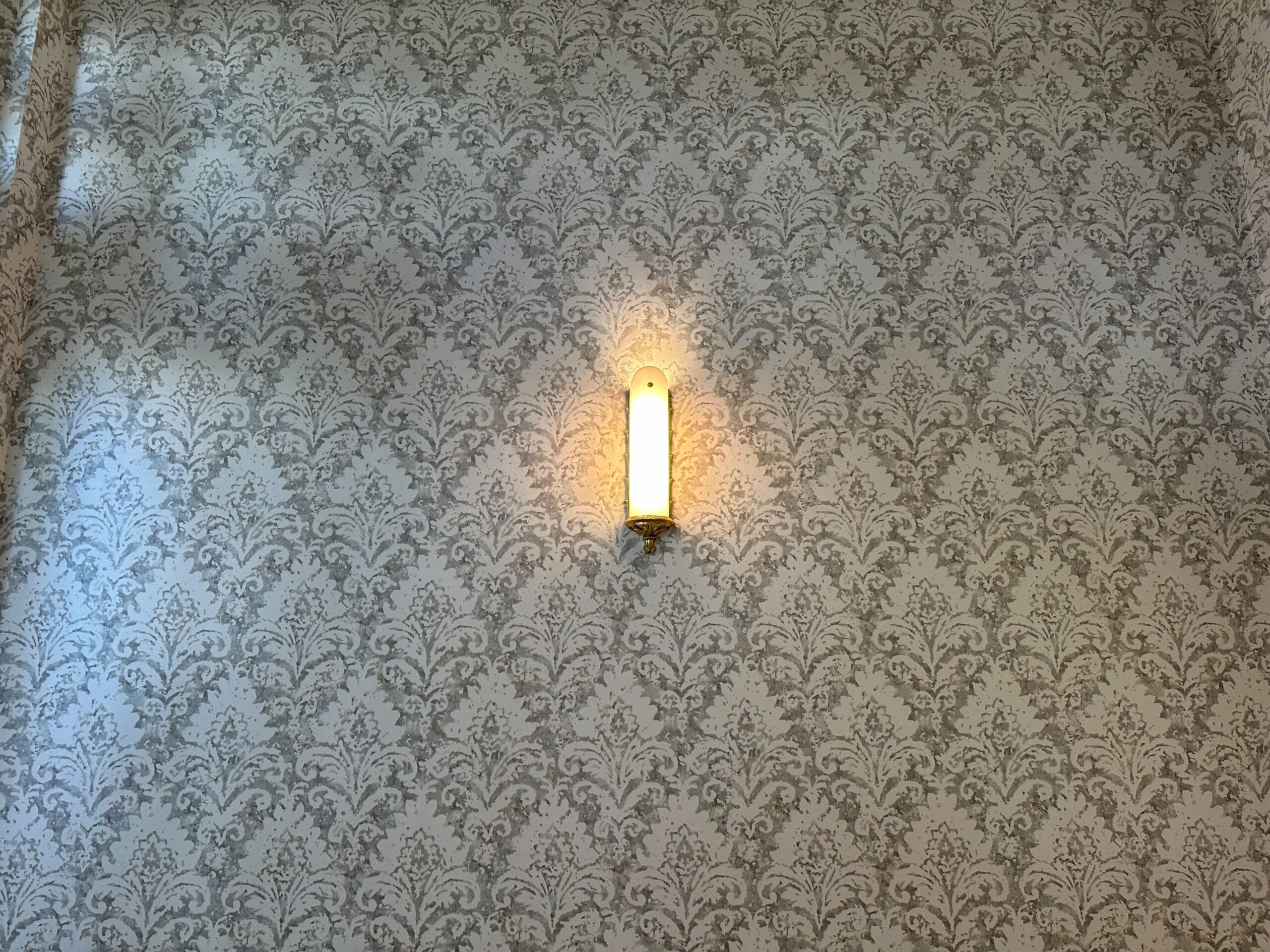 A wall sconce light illuminated on a wall with ornate, gray floral-patterned wallpaper.