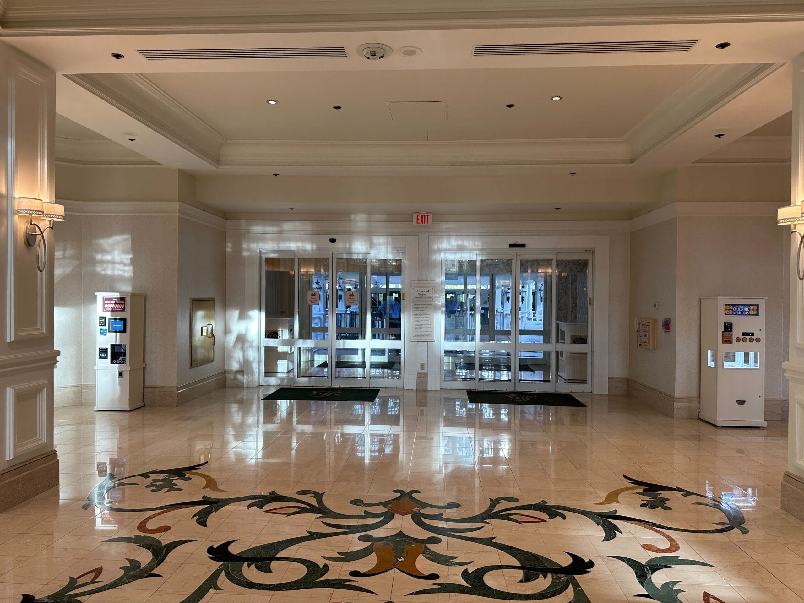Spacious hotel lobby with patterned tile floor, glass entrance doors, and two white kiosks on each side.