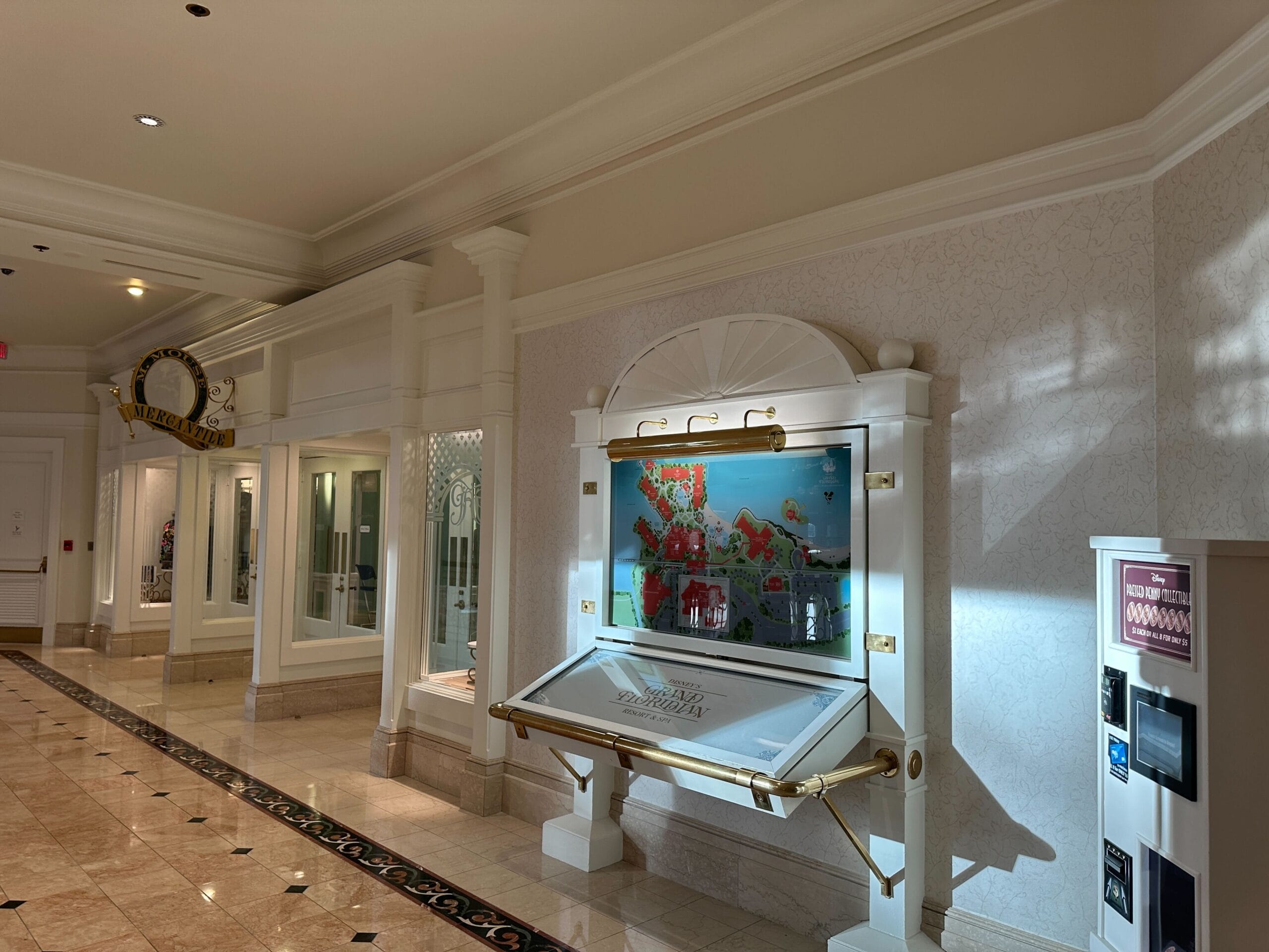 A bright hallway with a display case, wall art, marble floors, and storefronts on the left side.