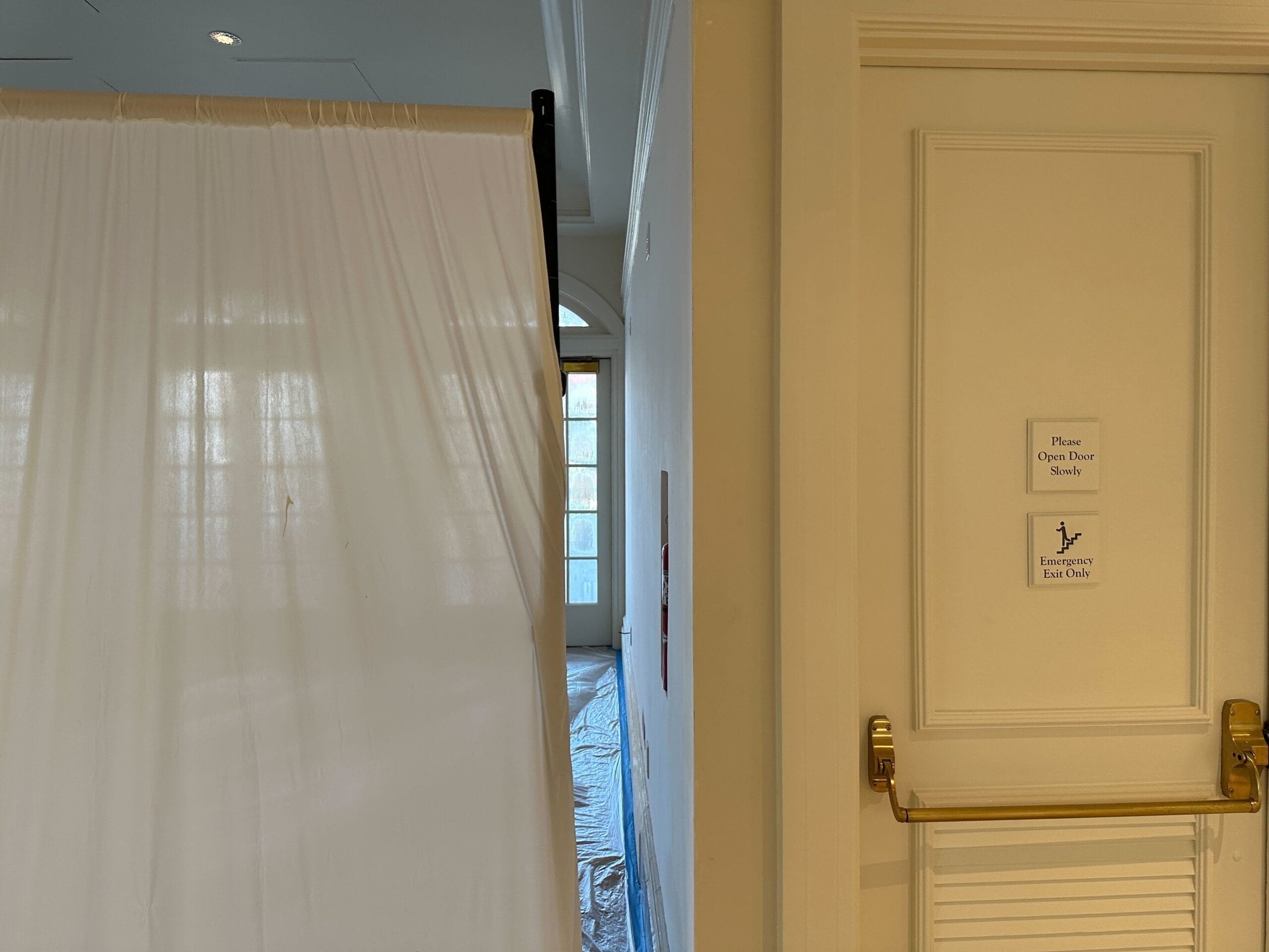 A hallway with a plastic sheet divider, a door with exit signs, and sunlight coming through windows at the end.
