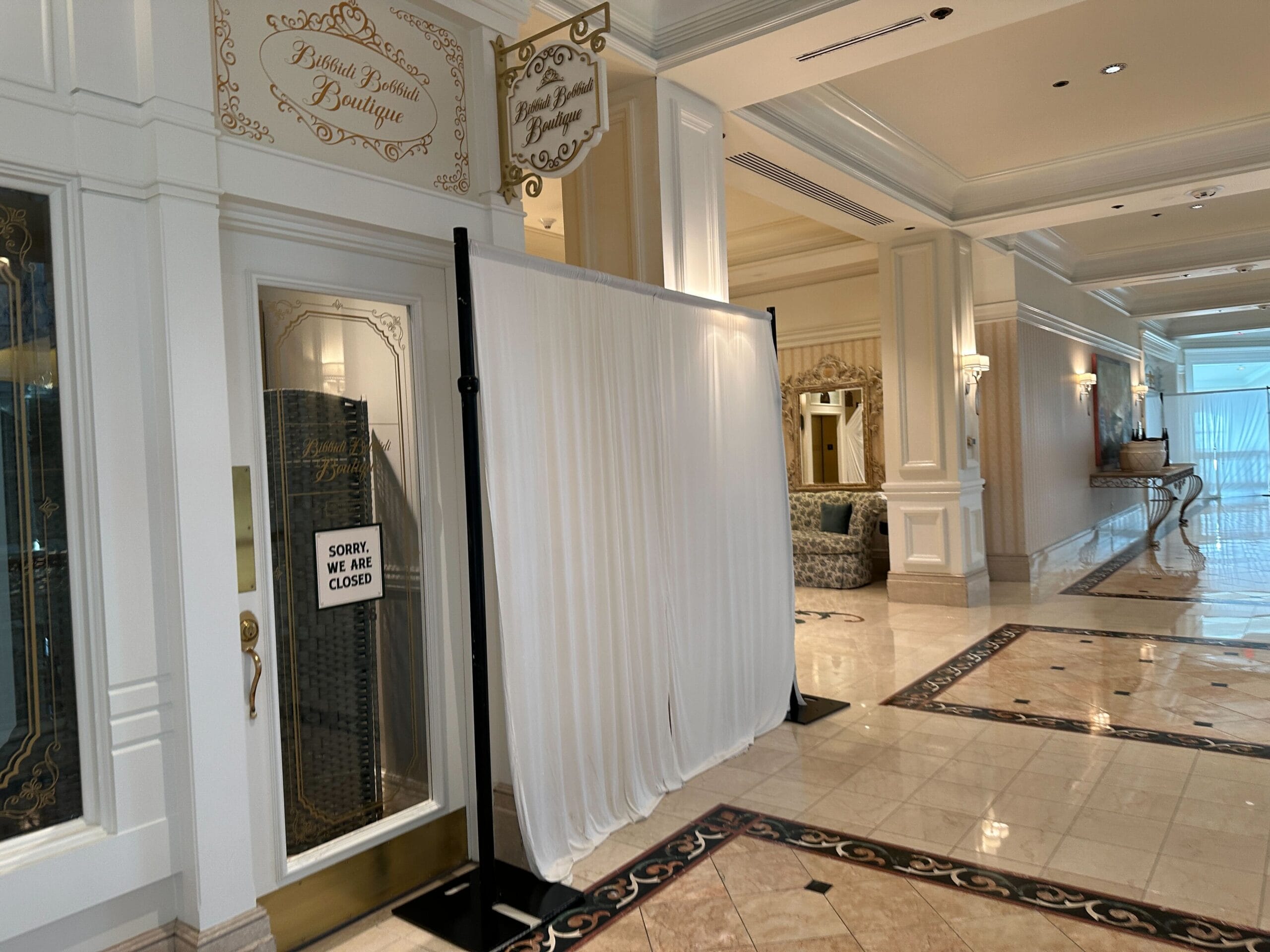 A boutique entrance with a "Sorry, we are closed" sign, blocked by white curtains in a marble hotel hallway.