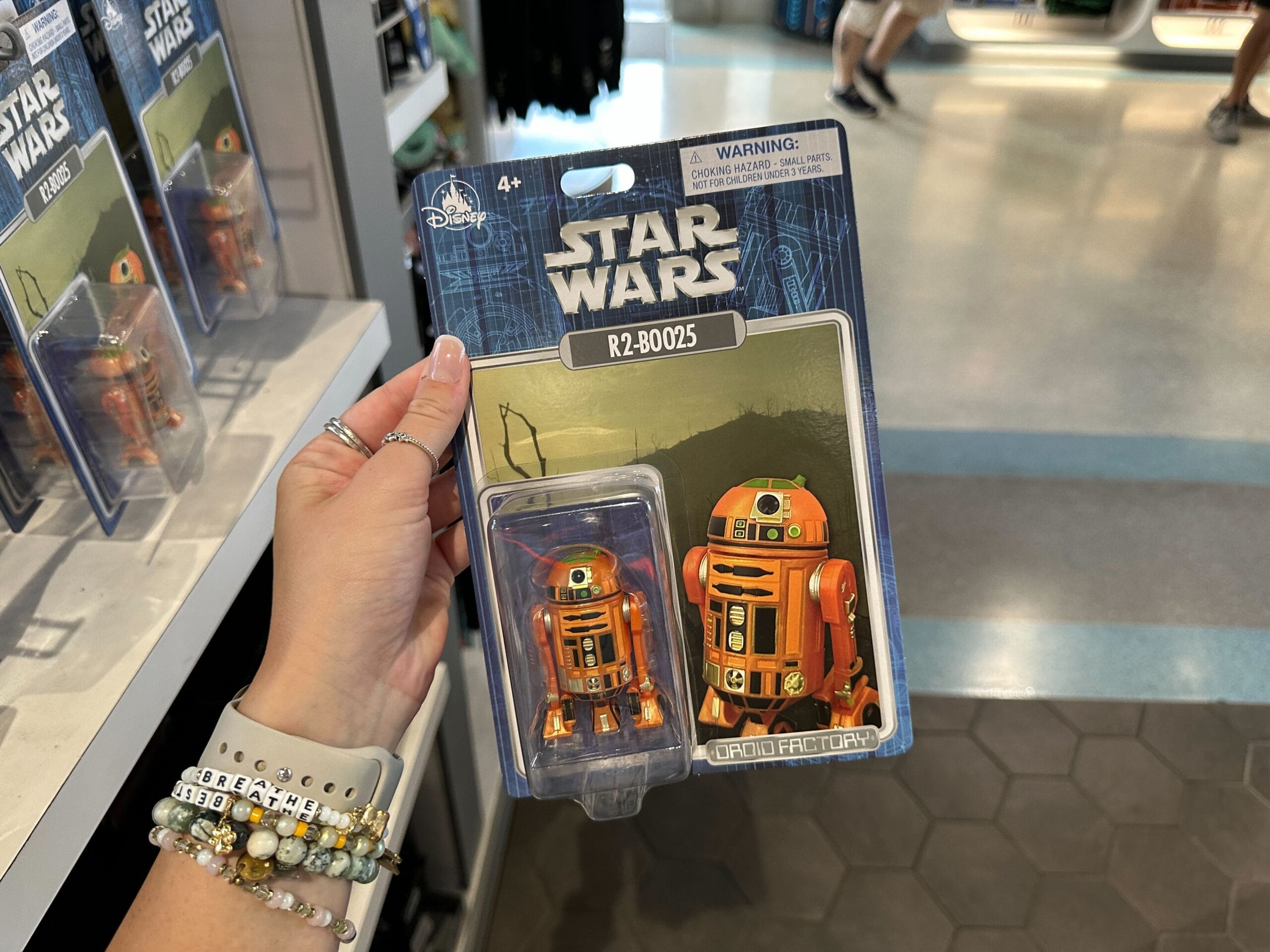Hand holding a packaged orange Star Wars R2-B0025 droid toy in a store with shelves and other toys in the background.