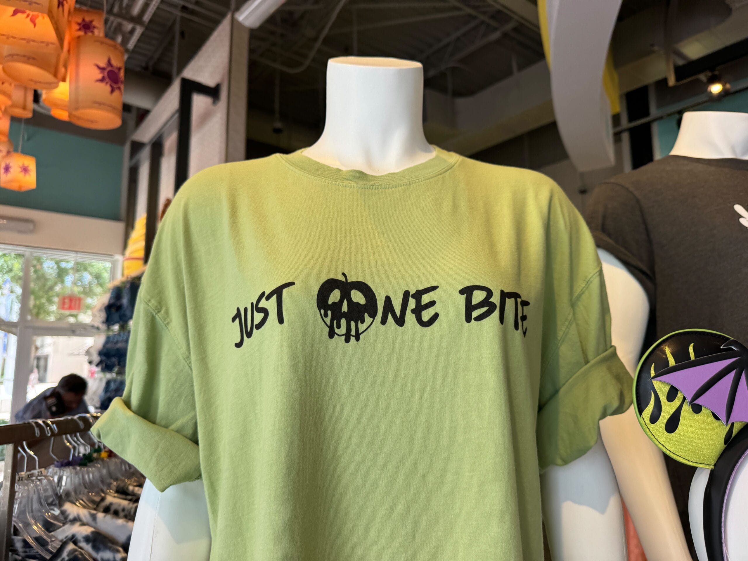A green t-shirt on a mannequin displays the text "JUST ONE BITE" with a poison apple graphic replacing the O.