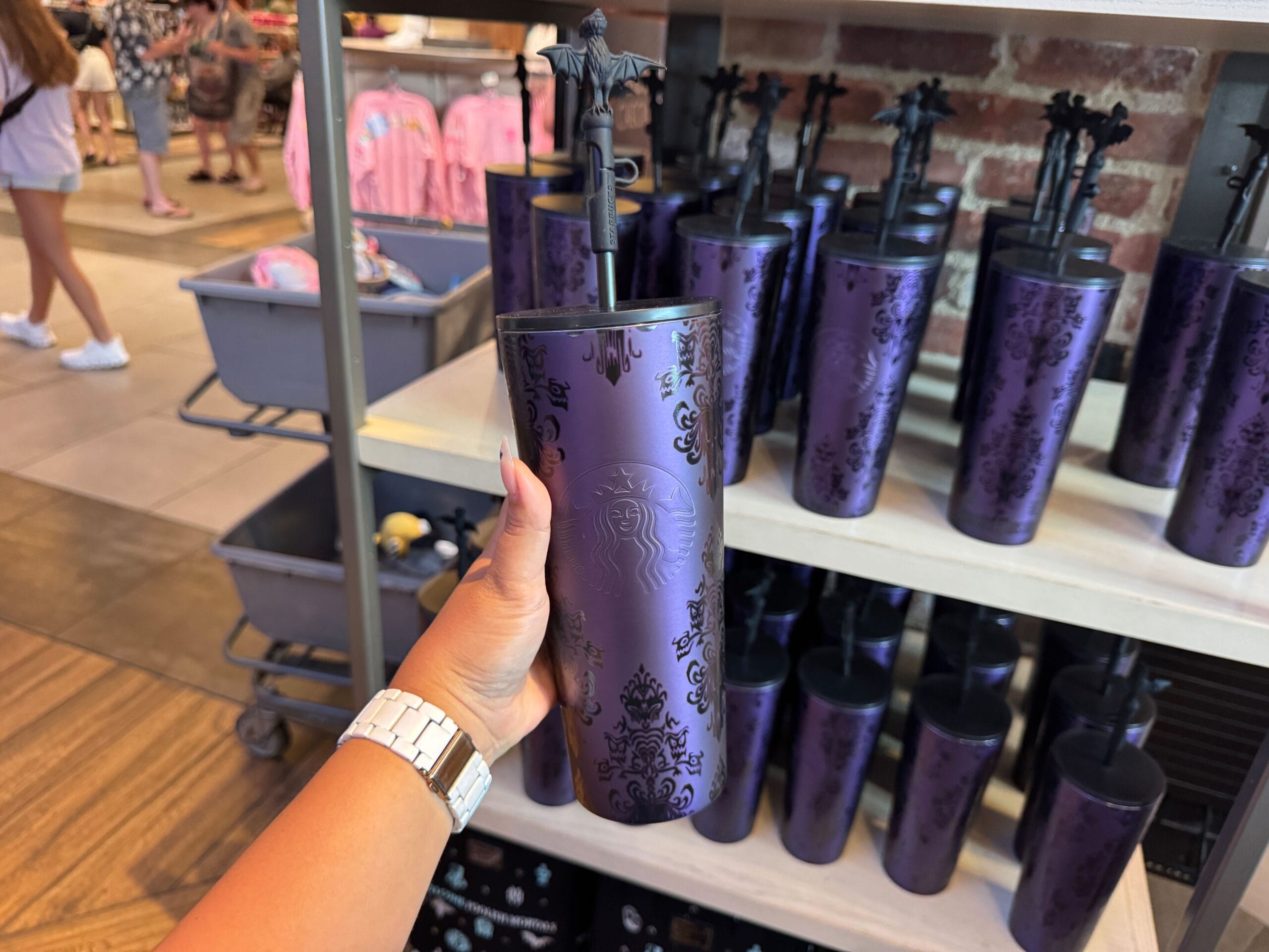 A hand holds a purple haunted mansion starbucks tumbler 2025 with a decorative straw in a store display filled with similar tumblers.