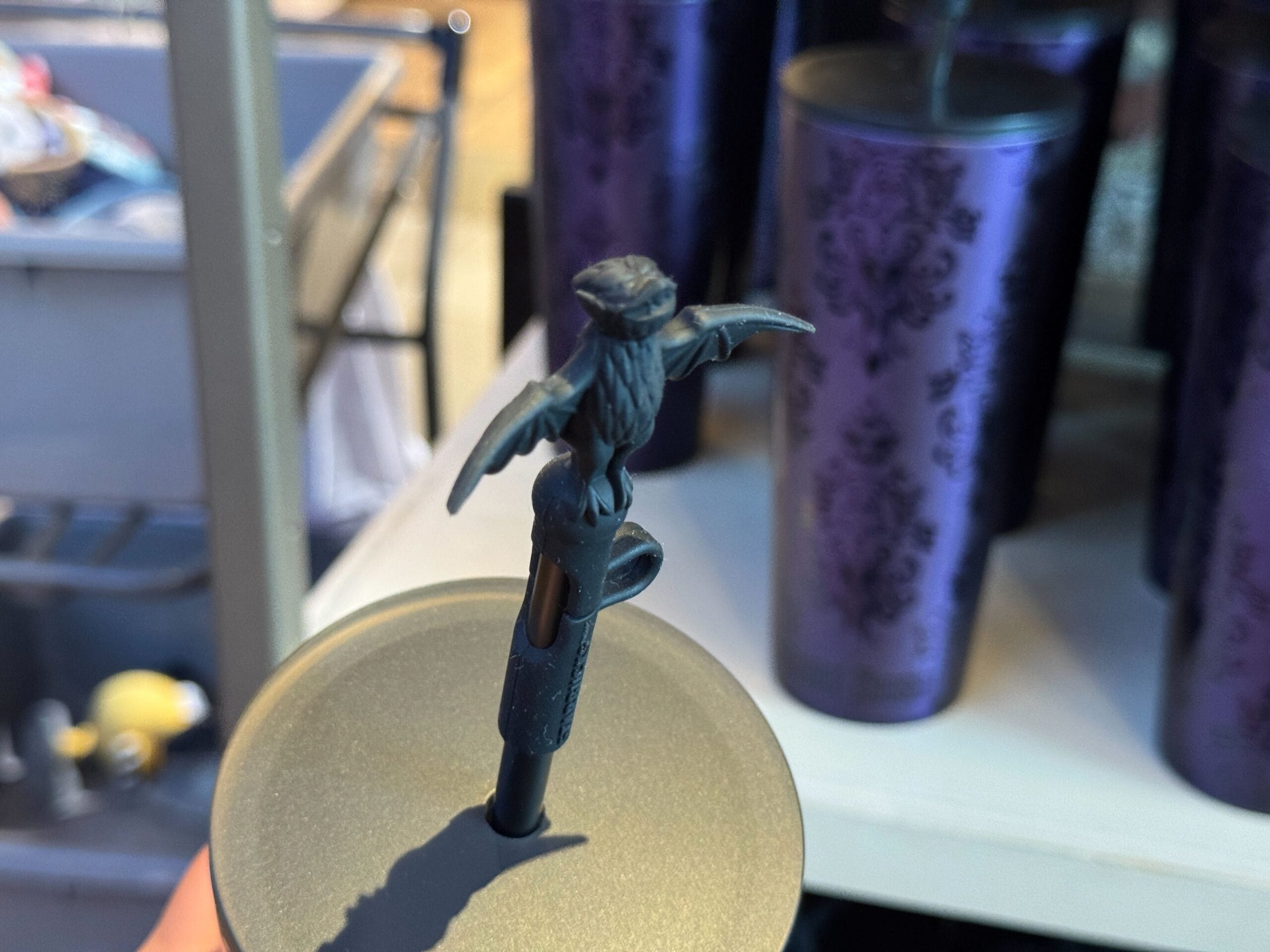 Close-up of a cup lid with a straw topped by a small, dark owl figurine; purple cups are in the background.