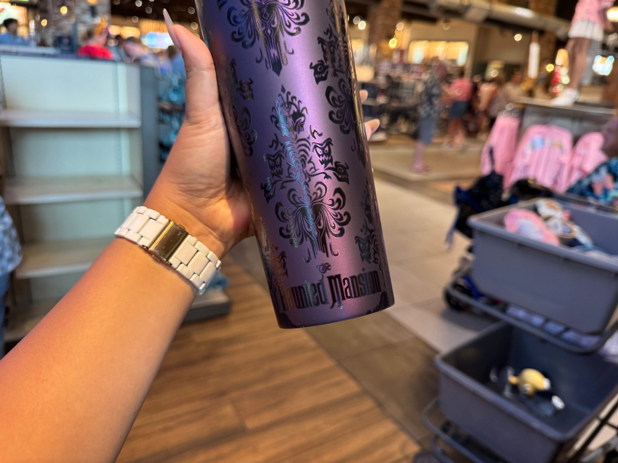 A hand holds a purple haunted mansion starbucks tumbler 2025 in a store with clothing racks and bins in the background.