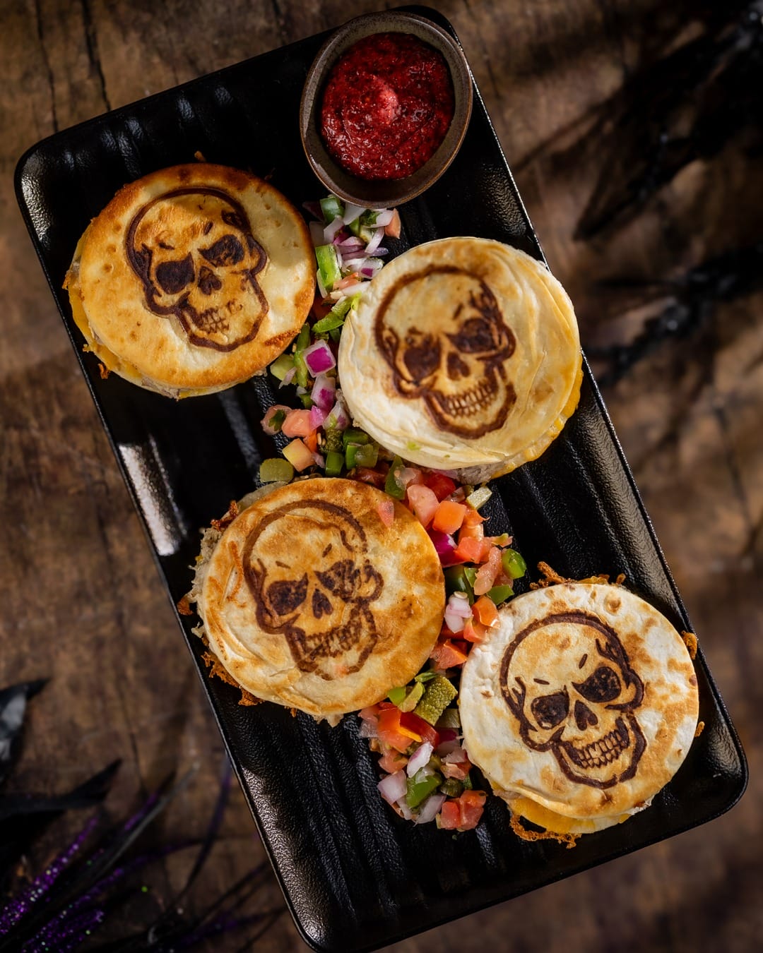 Jock Lindsey's Hangar Bar Halloween Four quesadillas with skull designs, served with salsa and colorful diced vegetables on a black rectangular plate.