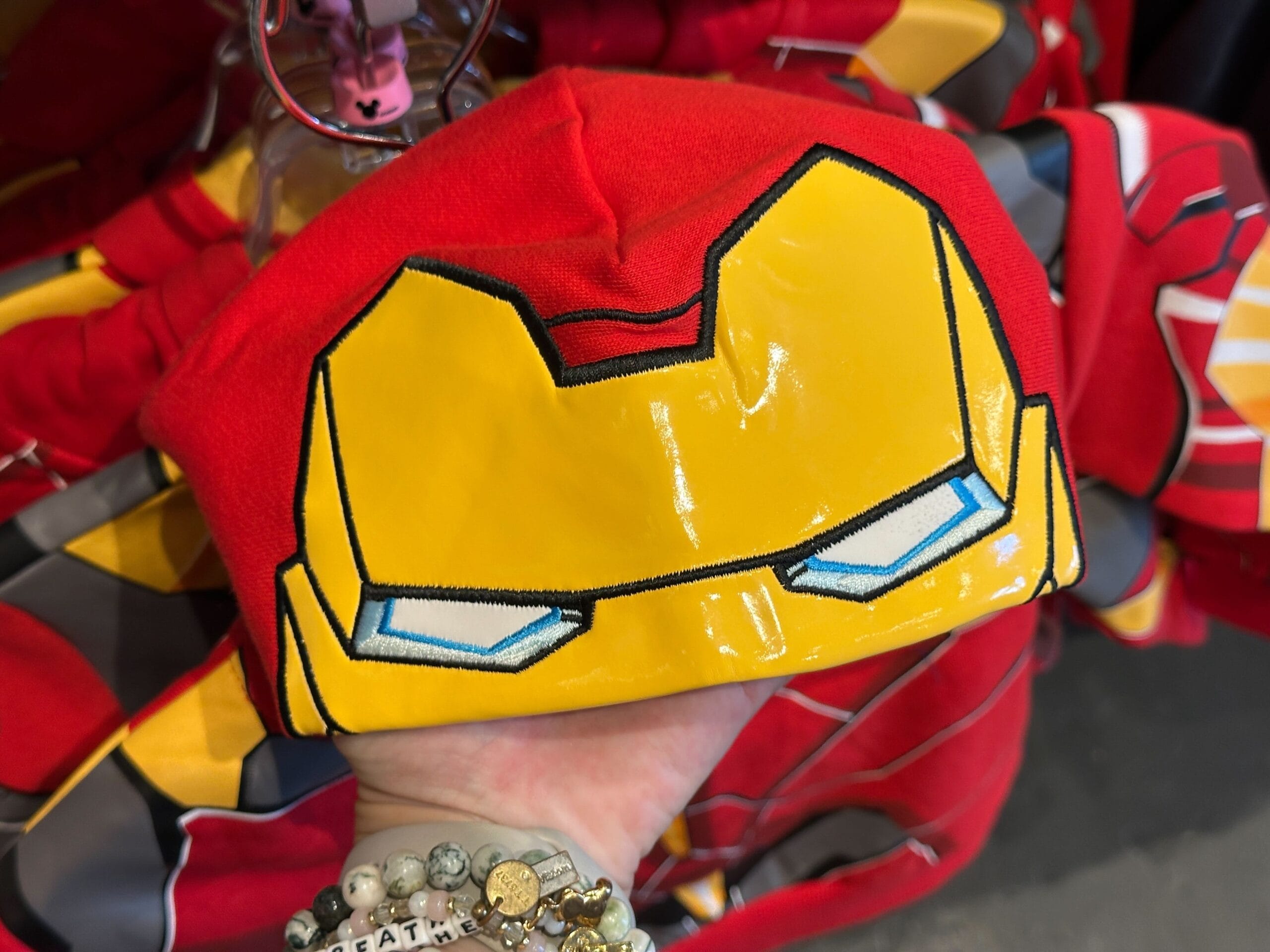 A hand holds a red beanie featuring a yellow Iron Man mask—perfect for fans of Youth Avengers Apparel.