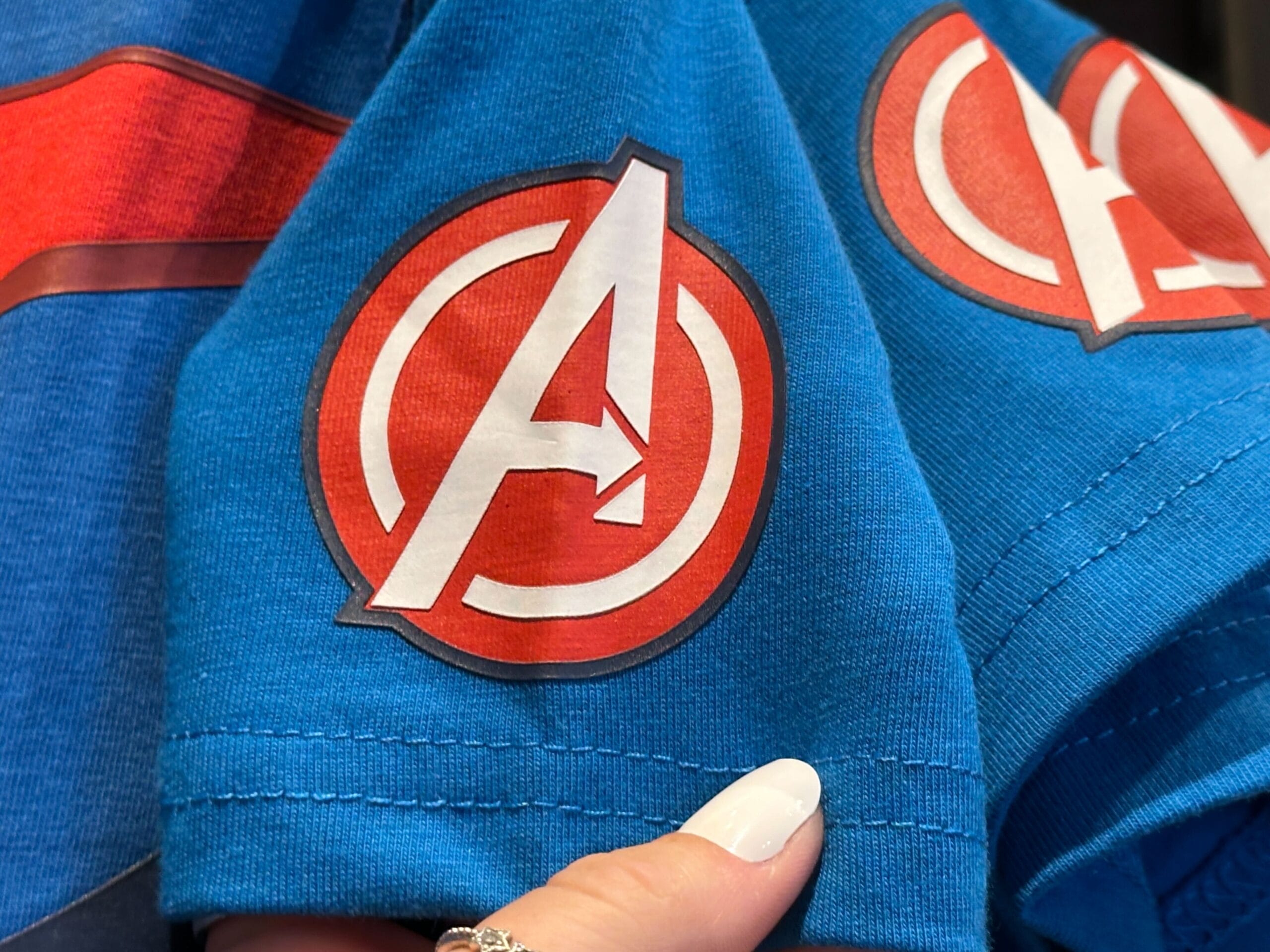 Close-up of a hand holding blue Youth Avengers Apparel with a red and white Avengers logo printed on it.