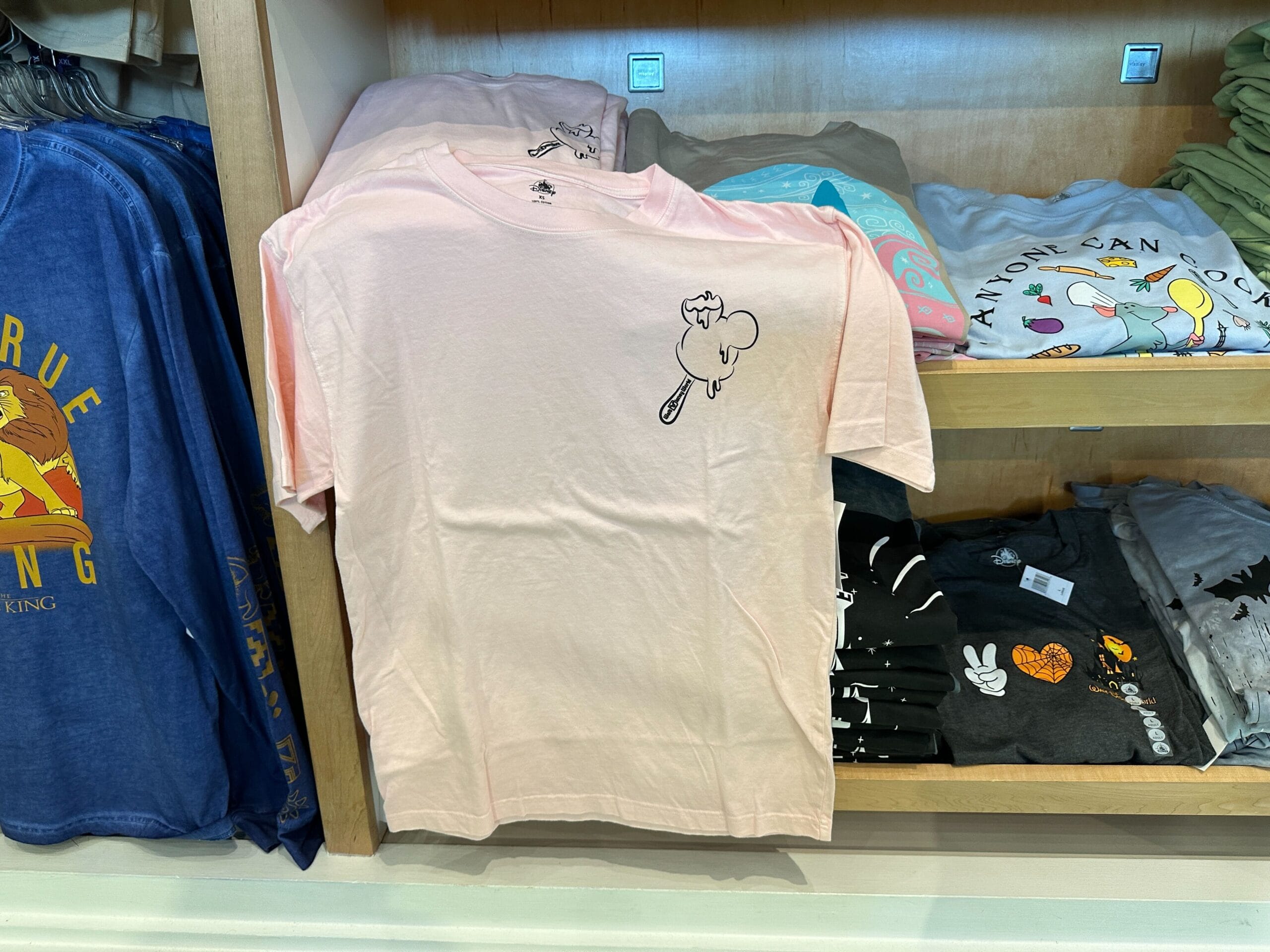 Light pink T-shirt with a small cat drawing holding a spoon printed on the upper left chest, displayed on a store shelf.