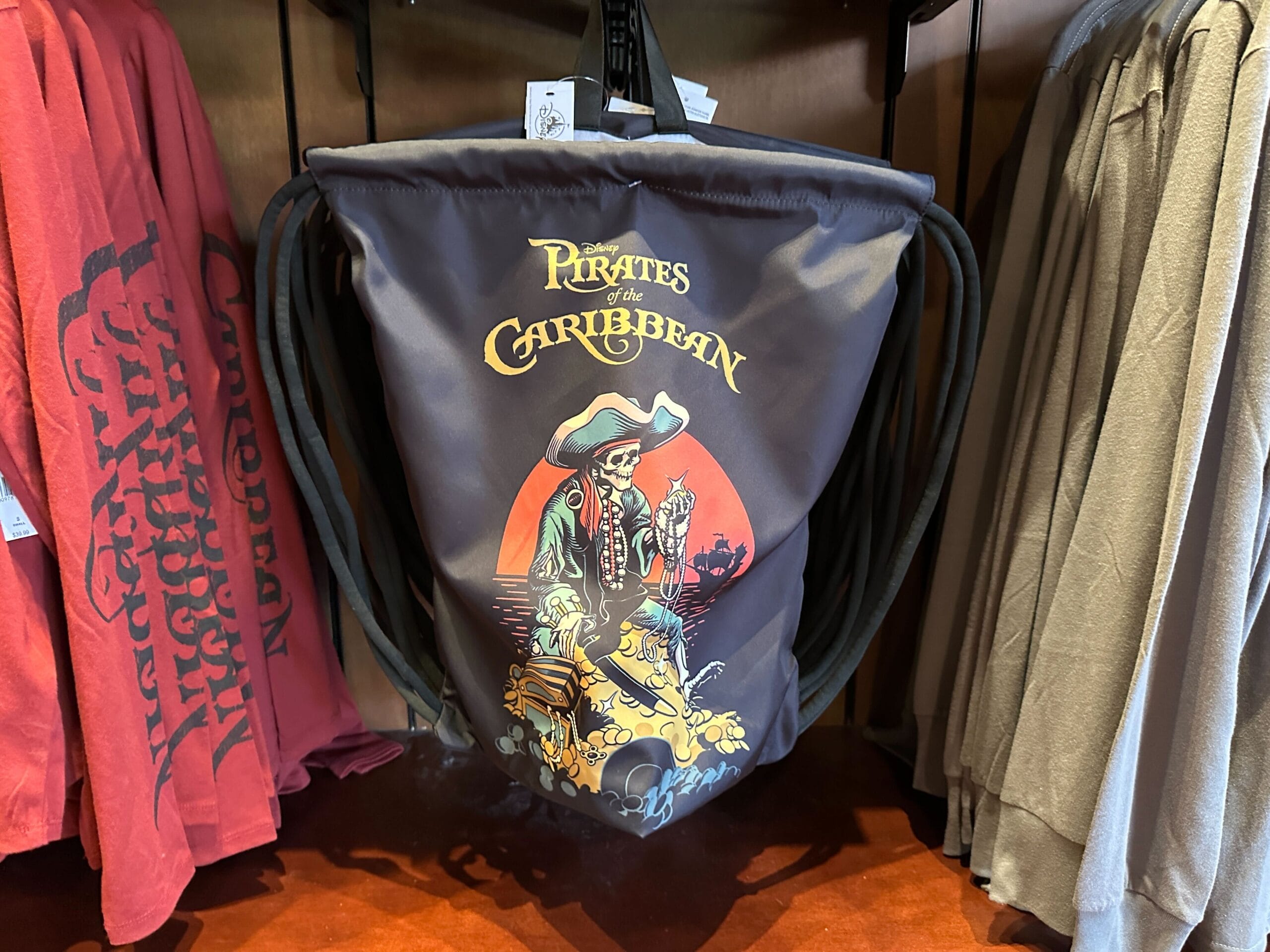 A drawstring bag with a Pirates of the Caribbean design featuring a pirate and treasure, hanging among shirts.