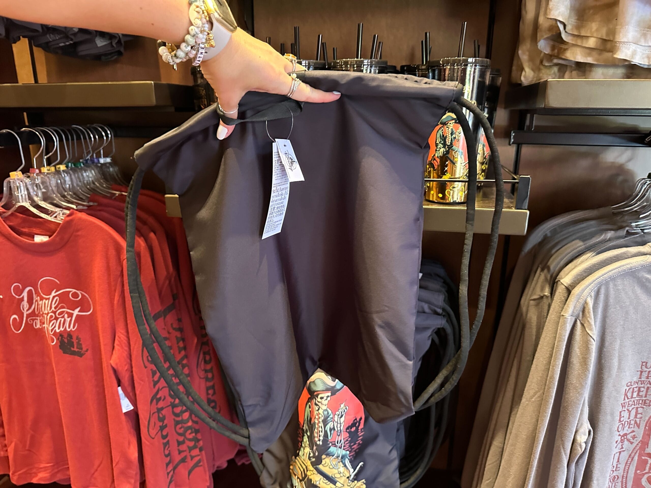 A person holds a black drawstring backpack in a store, surrounded by shirts and drink tumblers on shelves.