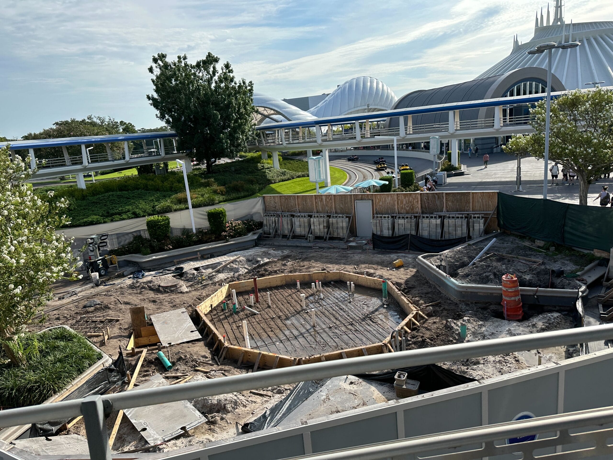 Construction site with a circular foundation and fencing in a futuristic theme park area under a blue sky.