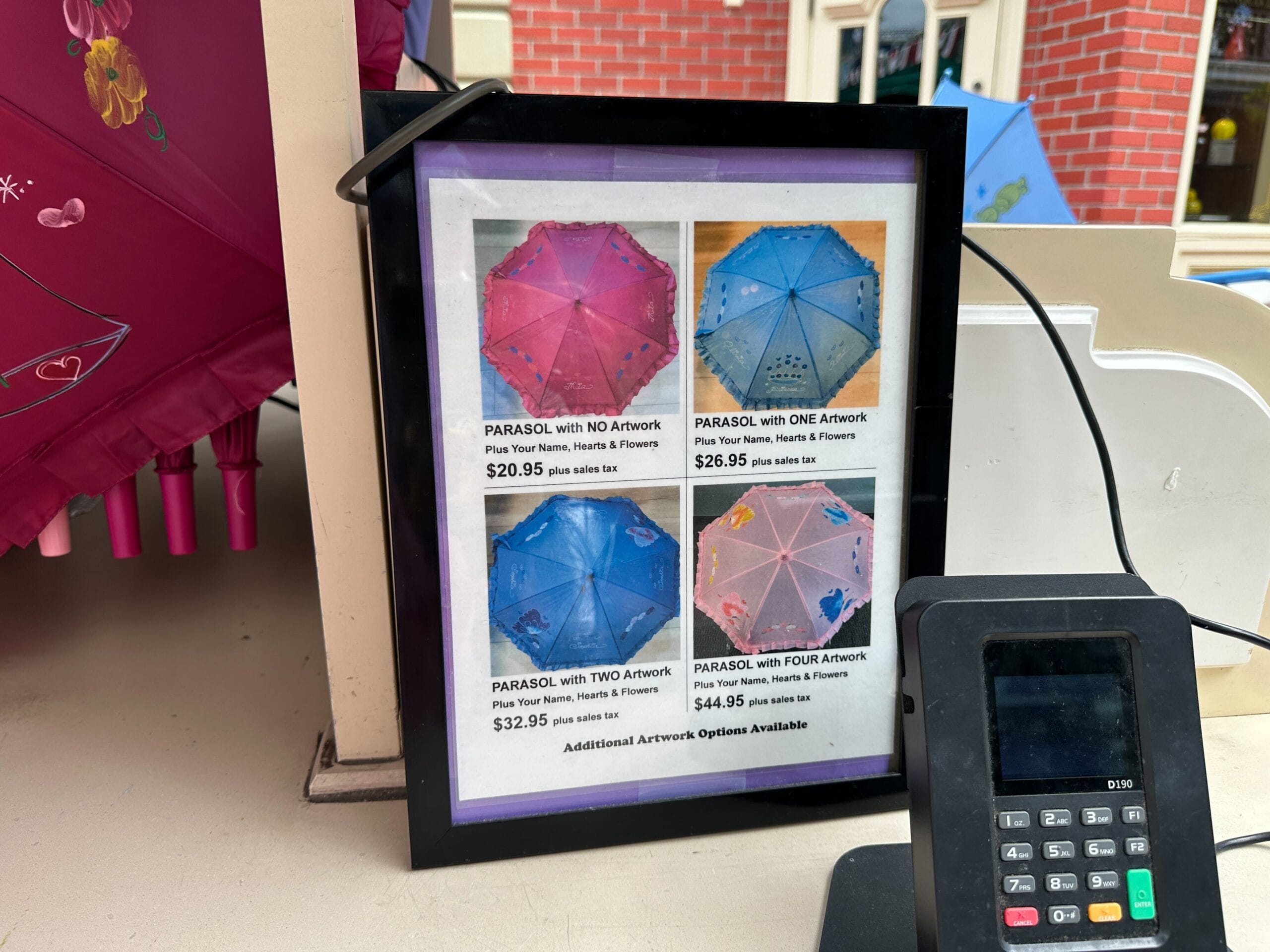 Framed sign showing parasol and caricature prices by artwork amount, next to a card reader on a counter.