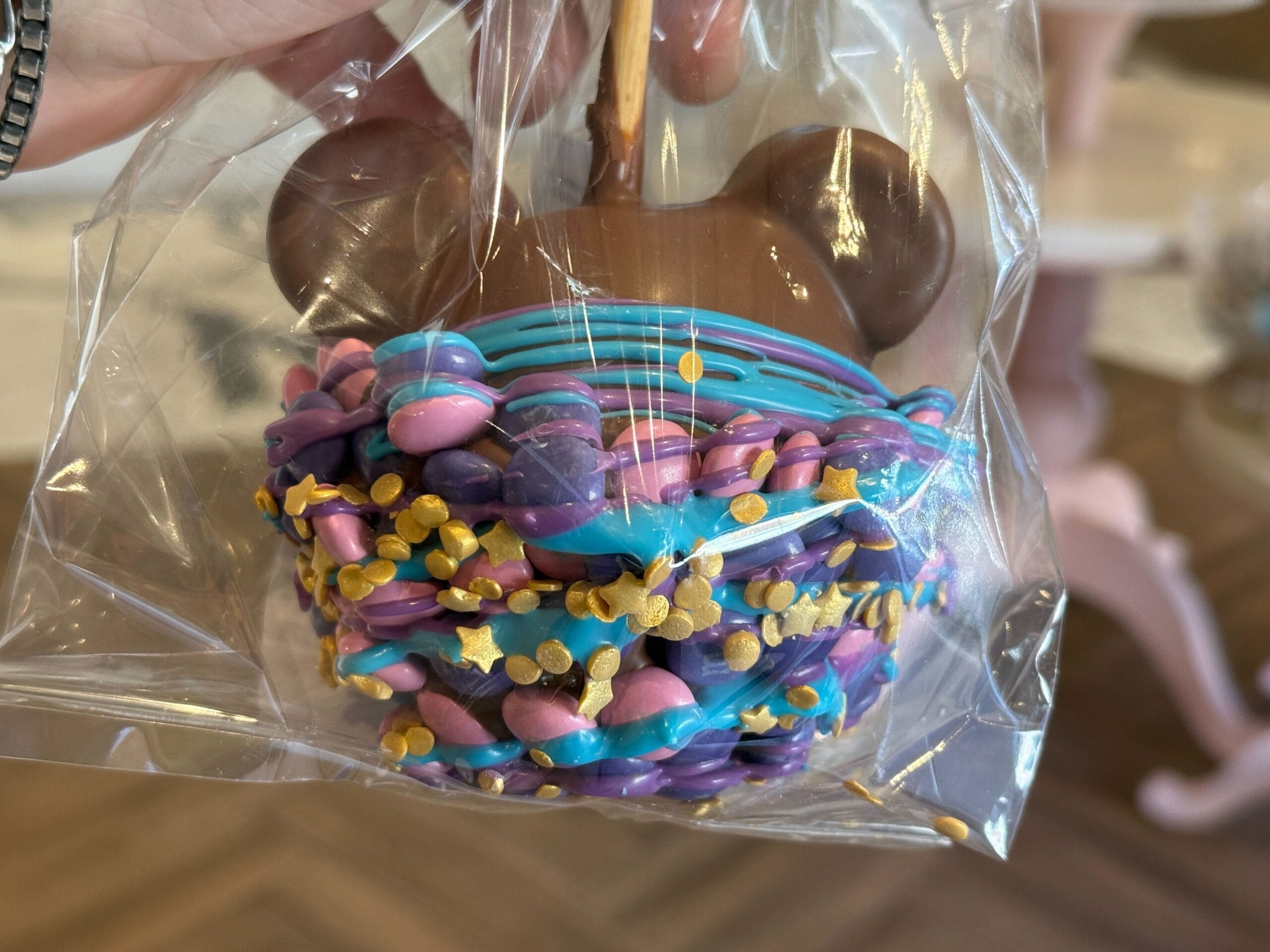 New Disney Parade Treats A hand holds a decorated candy apple, a sweet Magic Kingdom treat with chocolate ears, icing, and sprinkles in clear wrap.
