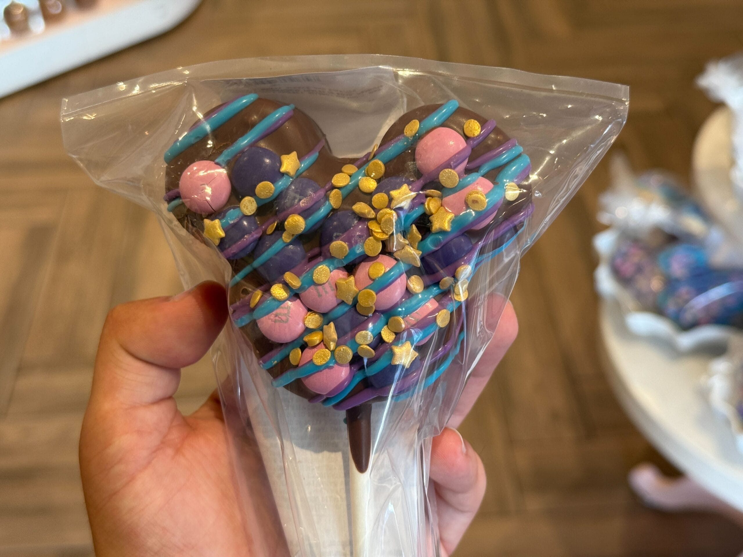 A hand holds a Mickey Mouse-shaped chocolate treat, a sweet Magic Kingdom snack wrapped in clear plastic with gold star sprinkles.