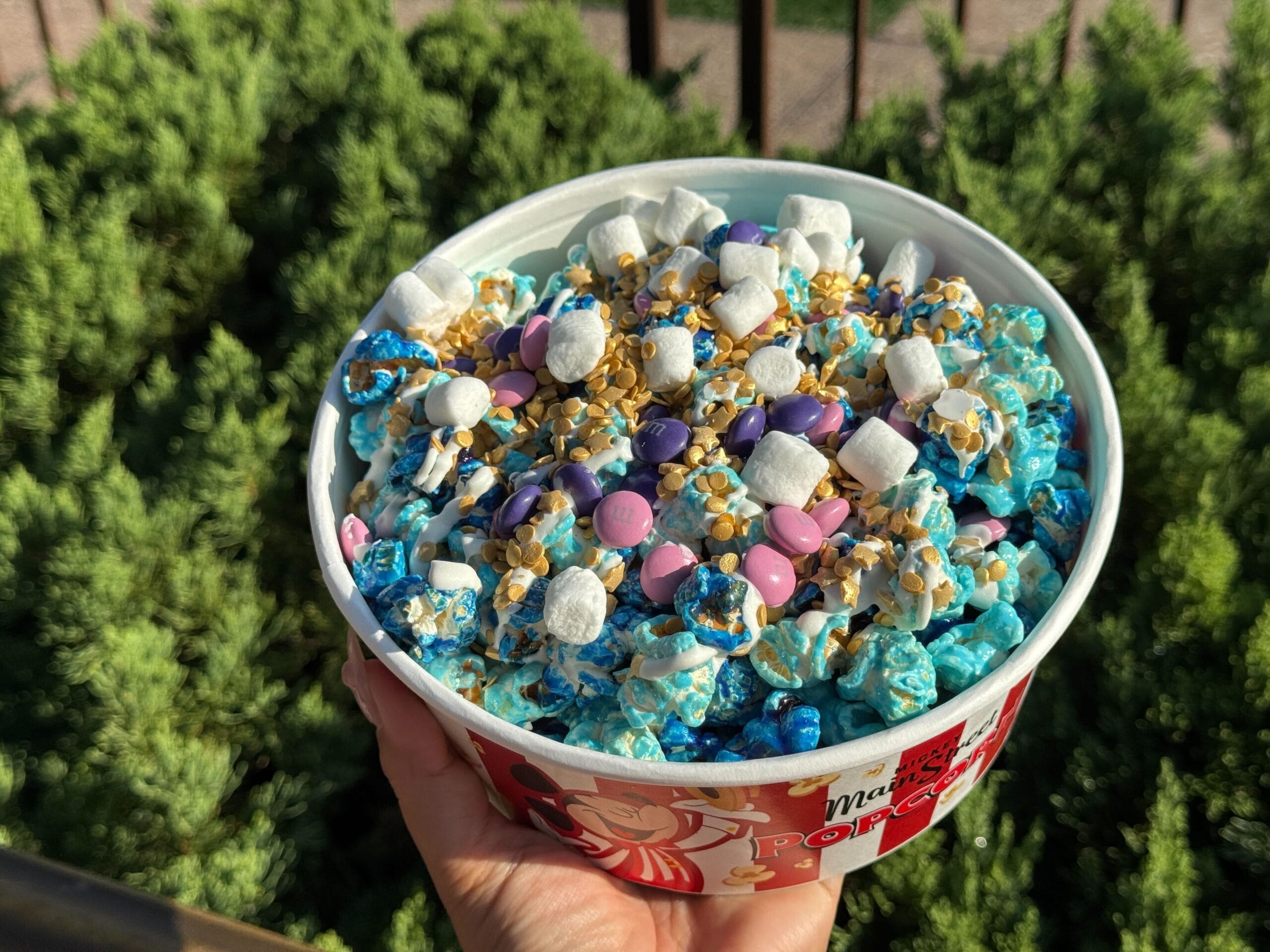 A hand holds a bowl of blue popcorn topped with candies, like festive Disney Starlight Parade snacks, with greenery behind.