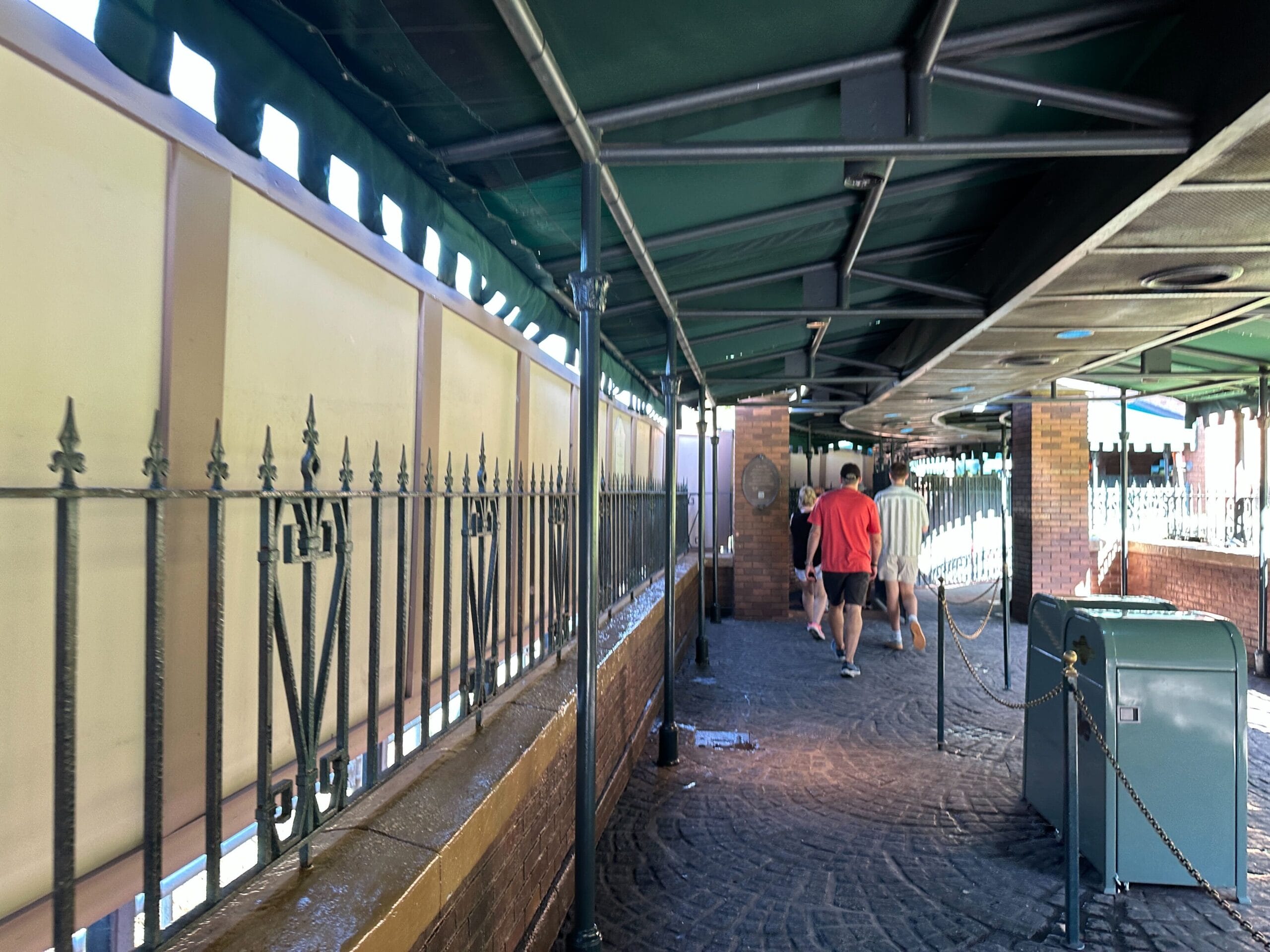 People walk down a shaded, fenced pathway near the Haunted Mansion with brick walls and a green trash can under a green awning.