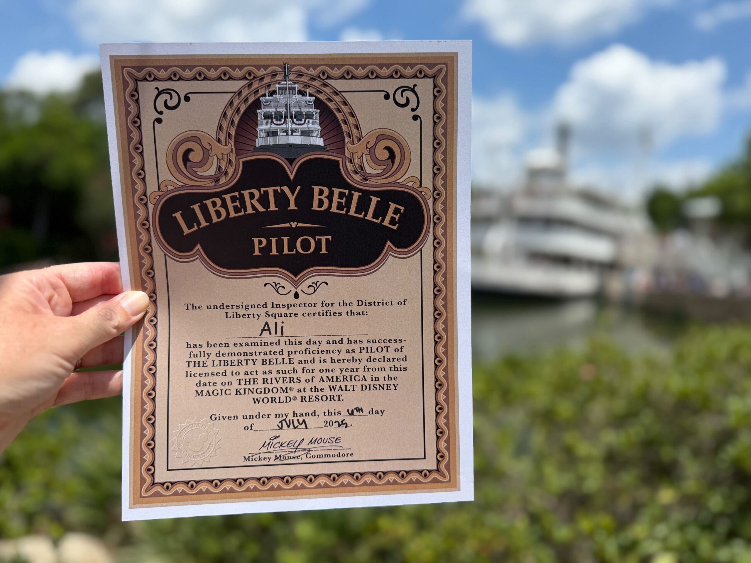 A hand holds a "Liberty Belle Pilot" certificate with the Liberty Square Riverboat and greenery blurred in the background.